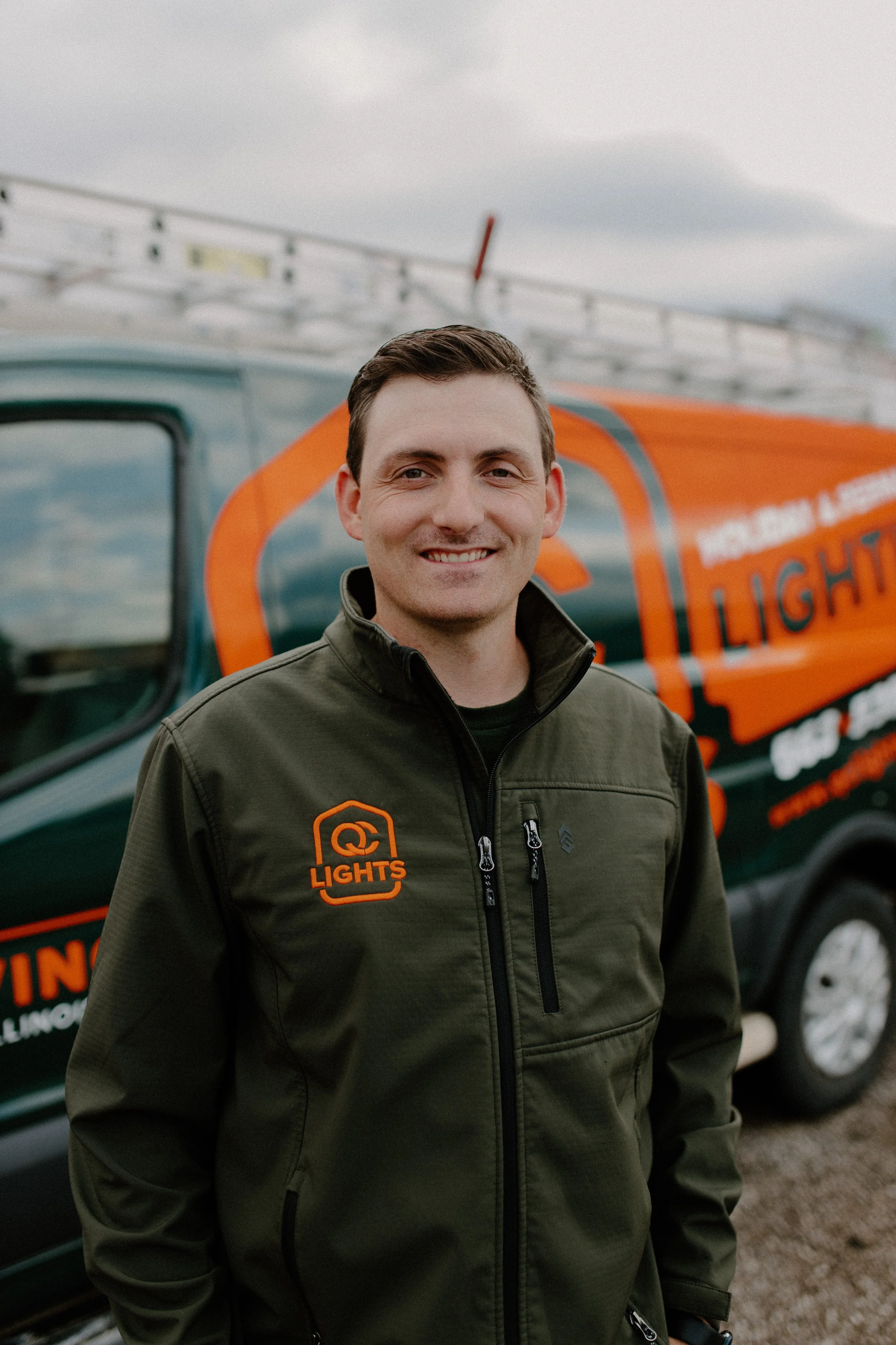 A man standing outdoors in front of a company branded vehicle, smiling, wearing a dark green jacket with an orange logo that reads 'Lights' on the chest.