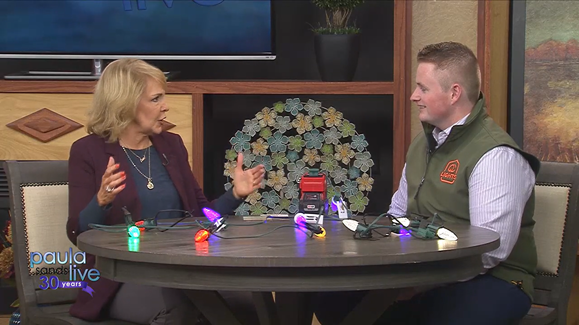 Two women engaged in a conversation during a television interview set, with colorful Christmas lights on the table between them. One woman, with blonde hair, is gesturing with her hands while talking, and the other woman, with short hair, is smiling. The backdrop includes a large screen, a potted plant, and wall art, suggesting a cozy, festive atmosphere.
