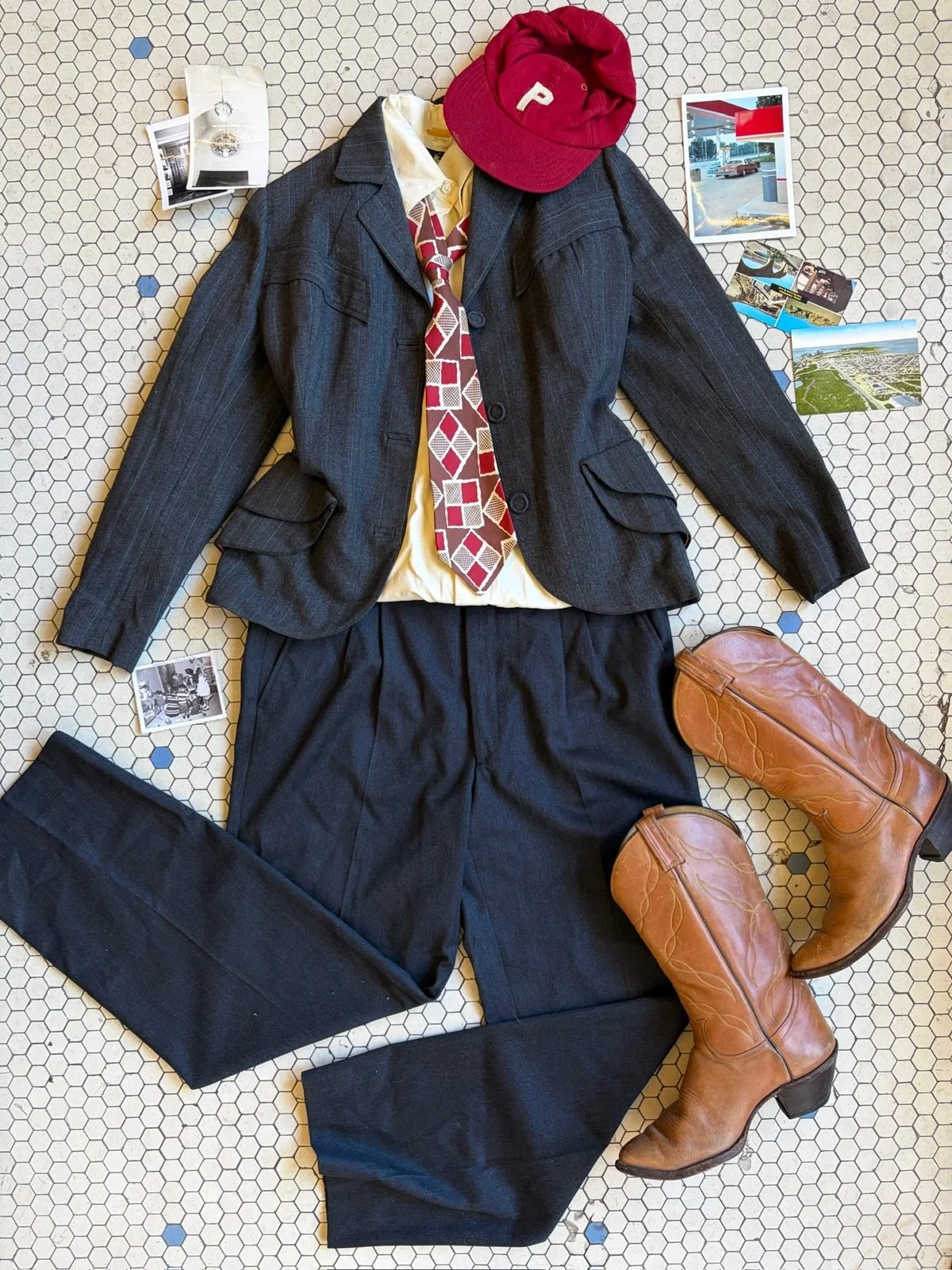 A schoolboy's outfit laid out on a tiled floor, including a dark gray pinstripe blazer, yellow shirt, patterned tie, dark gray trousers, brown cowboy boots, red baseball cap, and a brown leather bag. Surrounding items include photos and postcards.