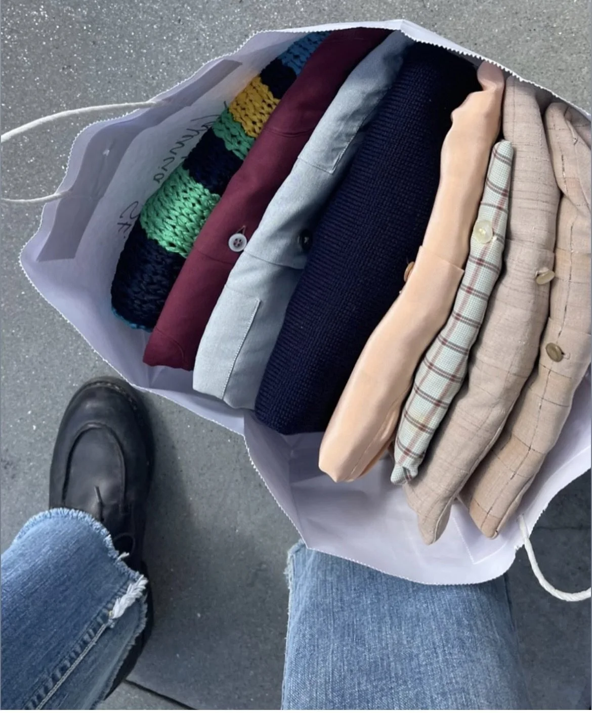 Open shopping bag containing folded clothing items, including a multicolored knit sweater, various jackets or blazers in different colors and fabrics. Foot of a person wearing a black boot and blue jeans is visible in the bottom left corner.