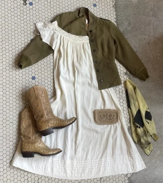 A vintage-style outfit laid out on a white tiled floor, including a white dress with lace details, a green jacket, a beige purse, cowboy boots, and a yellow scarf with black accents.