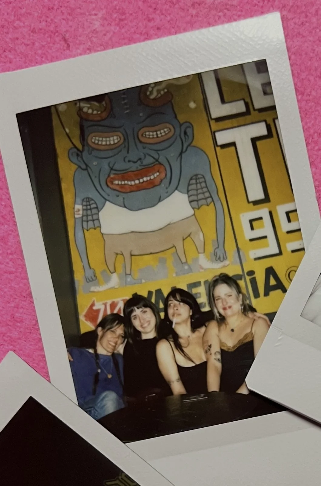 Group of four women posing in front of a mural featuring a cartoonish, exaggerated character with multiple eyes, horns, and a humanoid body.