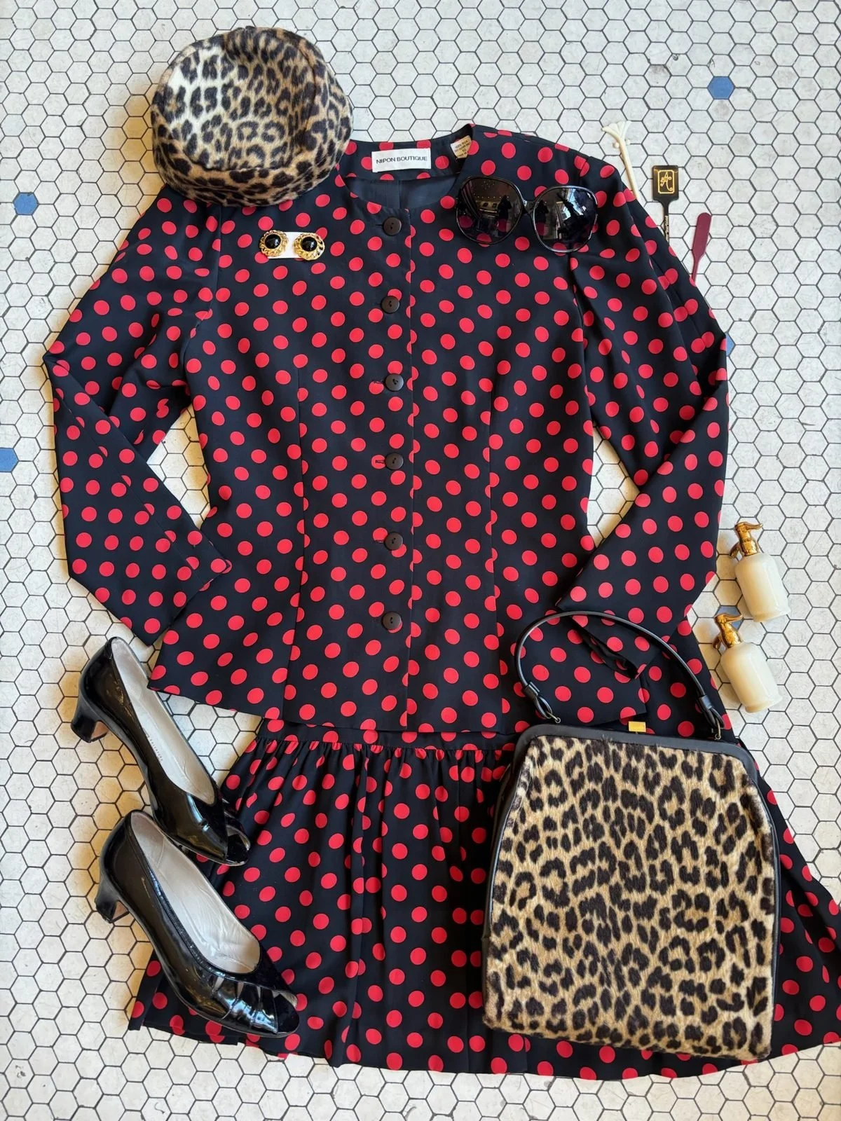 Flat lay of a polka dot dress, leopard print purse, black high heels, black sunglasses, and small bottles of lotion on a honeycomb tile floor.