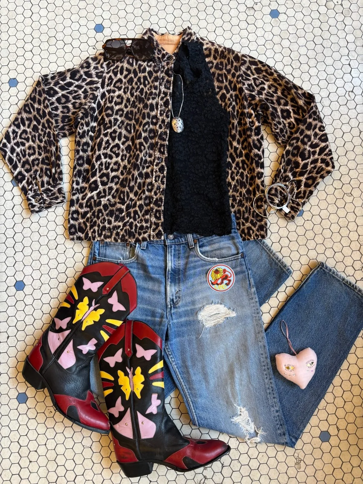 Leopard print jacket, black lace top, ripped jeans with a patch, colorful butterfly cowboy boots, black sunglasses, necklace, bracelet, and a pink heart-shaped plush toy with eyes on a honeycomb tile floor.