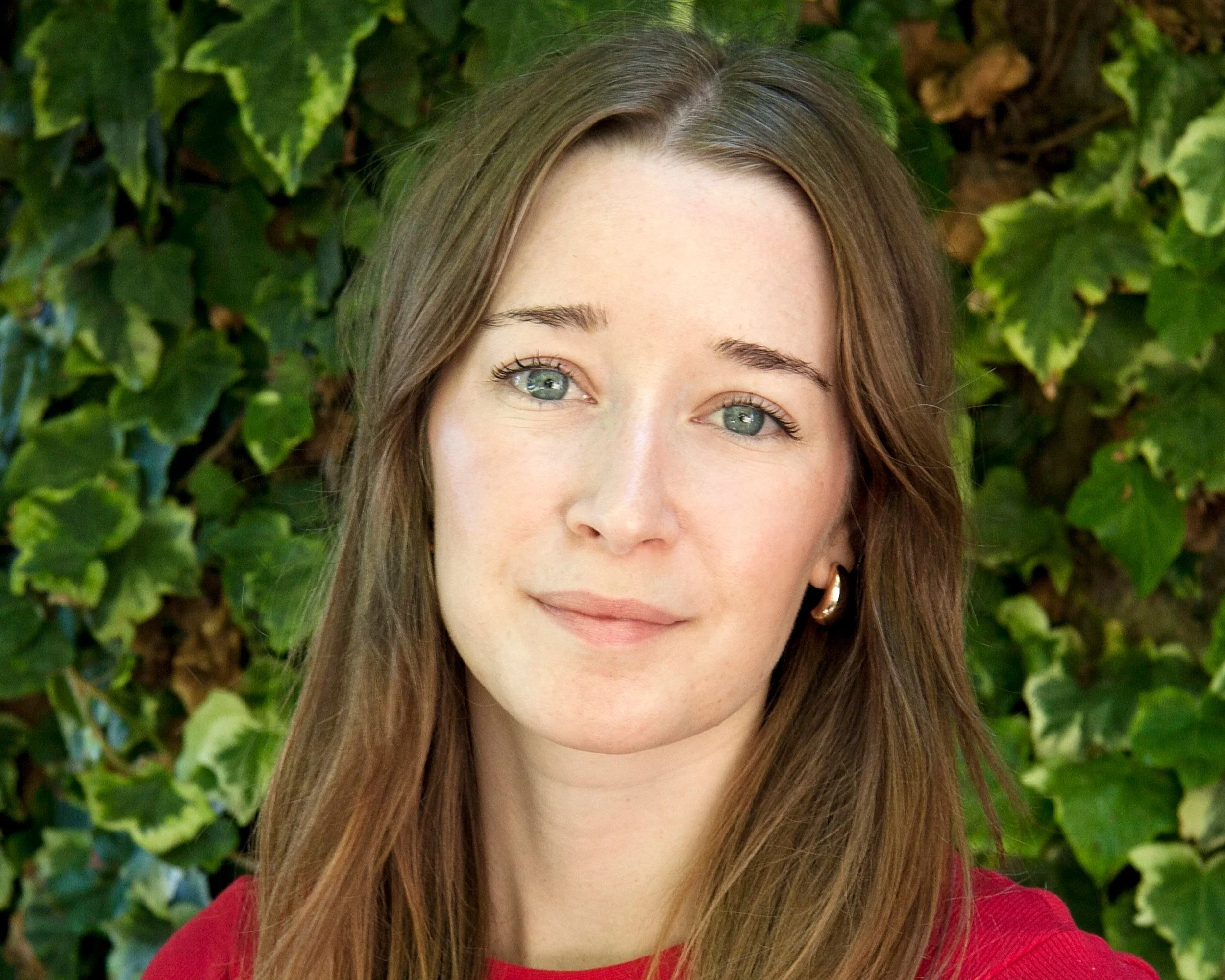 Portrait of a young woman with long brown hair, light skin, wearing a red top and hoop earrings, standing outdoors with green ivy leaves in the background.