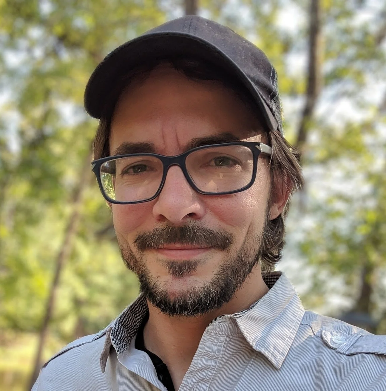 A selfie of a man wearing glasses and a cap. With trees in the background. 