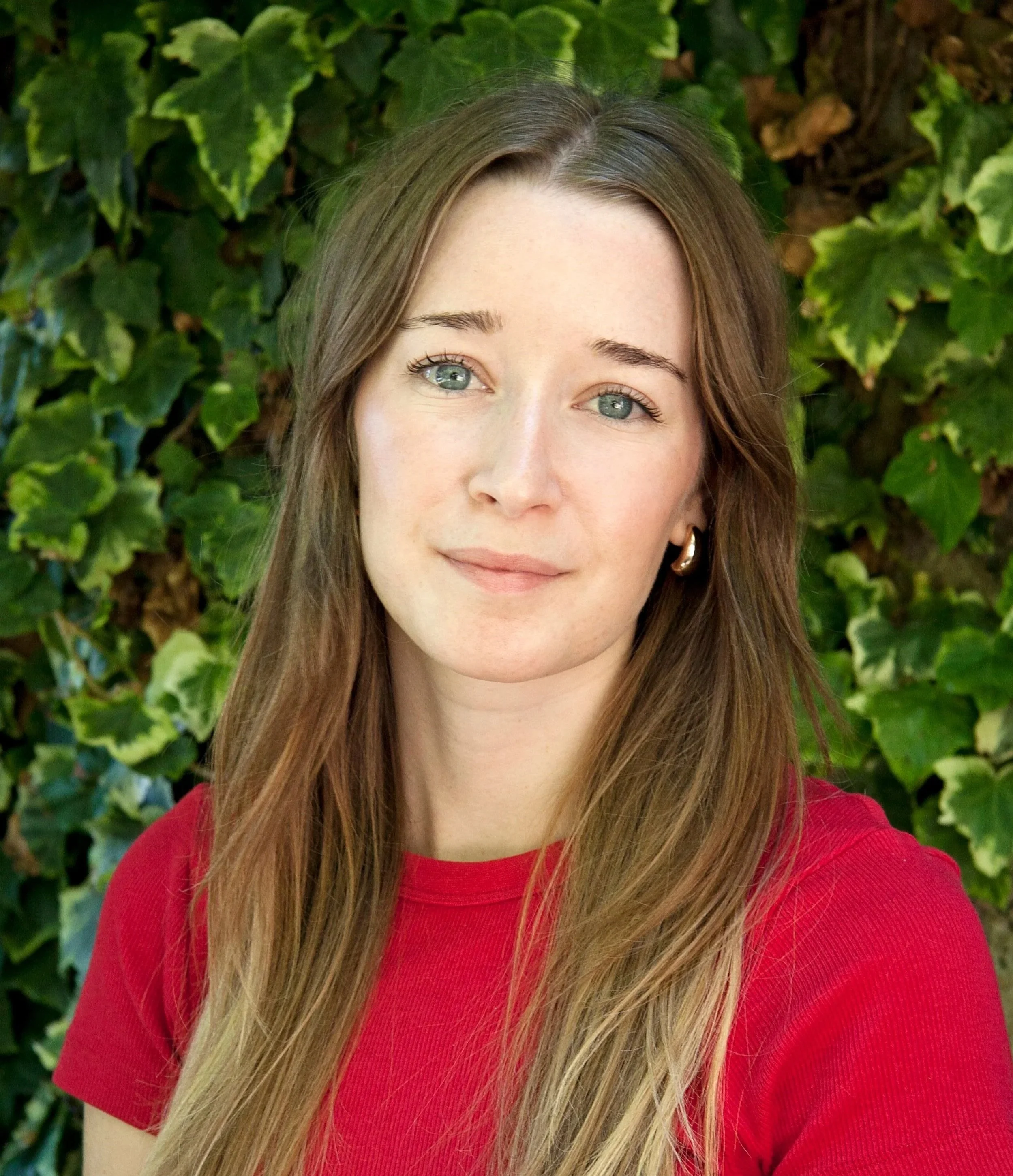Portrait of a young woman with long brown hair, light skin, wearing a red top and hoop earrings, standing outdoors with green ivy leaves in the background.