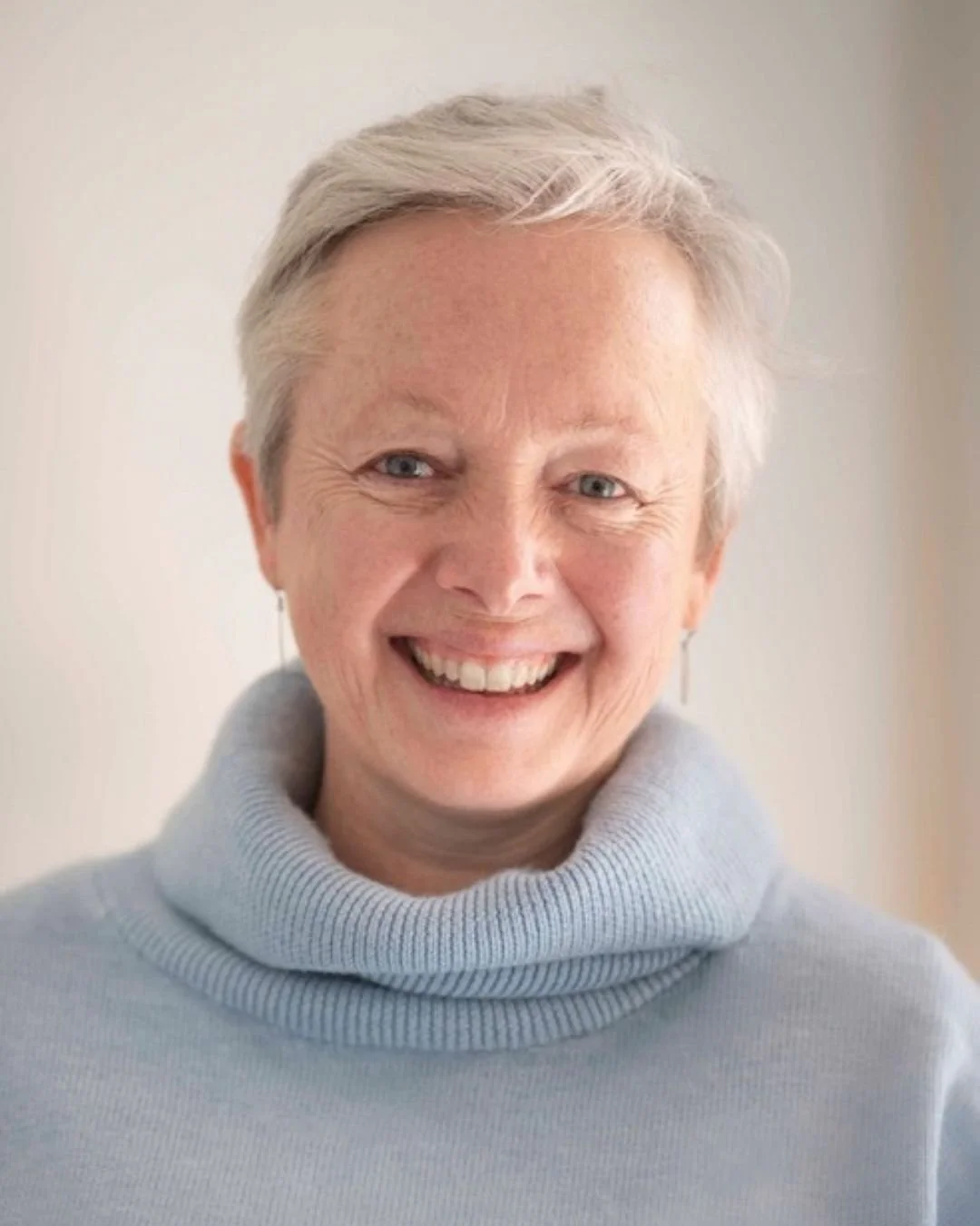 A smiling woman with short gray hair wearing a light gray turtleneck sweater and earrings.