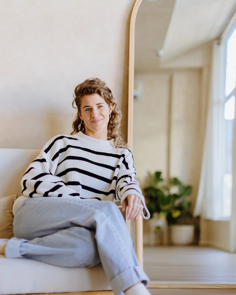 A smiling woman with curly hair sitting on a beige couch in a bright room with cream-colored walls, a wooden mirror, and large window with white curtains, dressed in a white and black striped sweater and light gray pants.