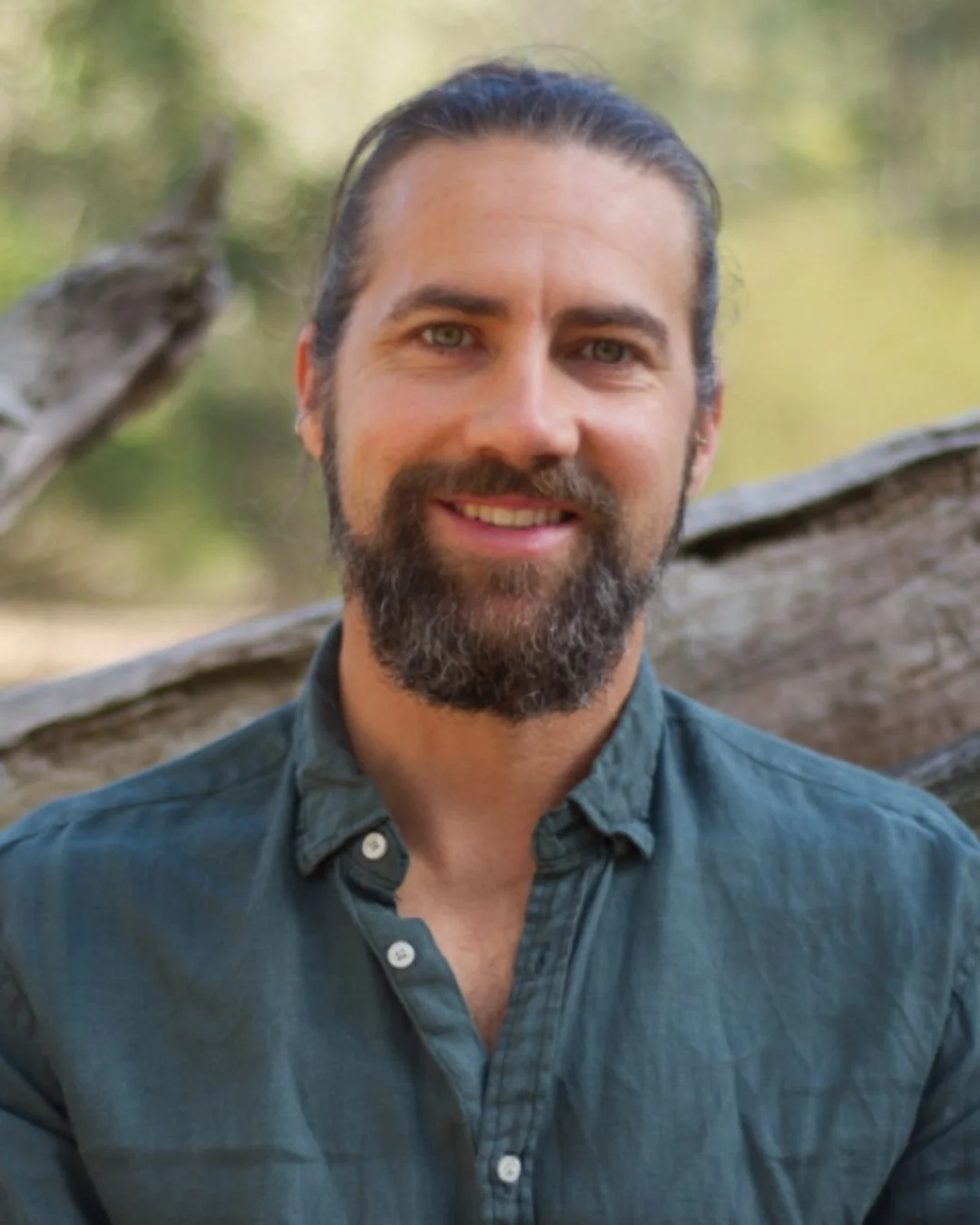 A man with long dark hair and a beard, smiling, outdoors in front of a fallen tree and green foliage.