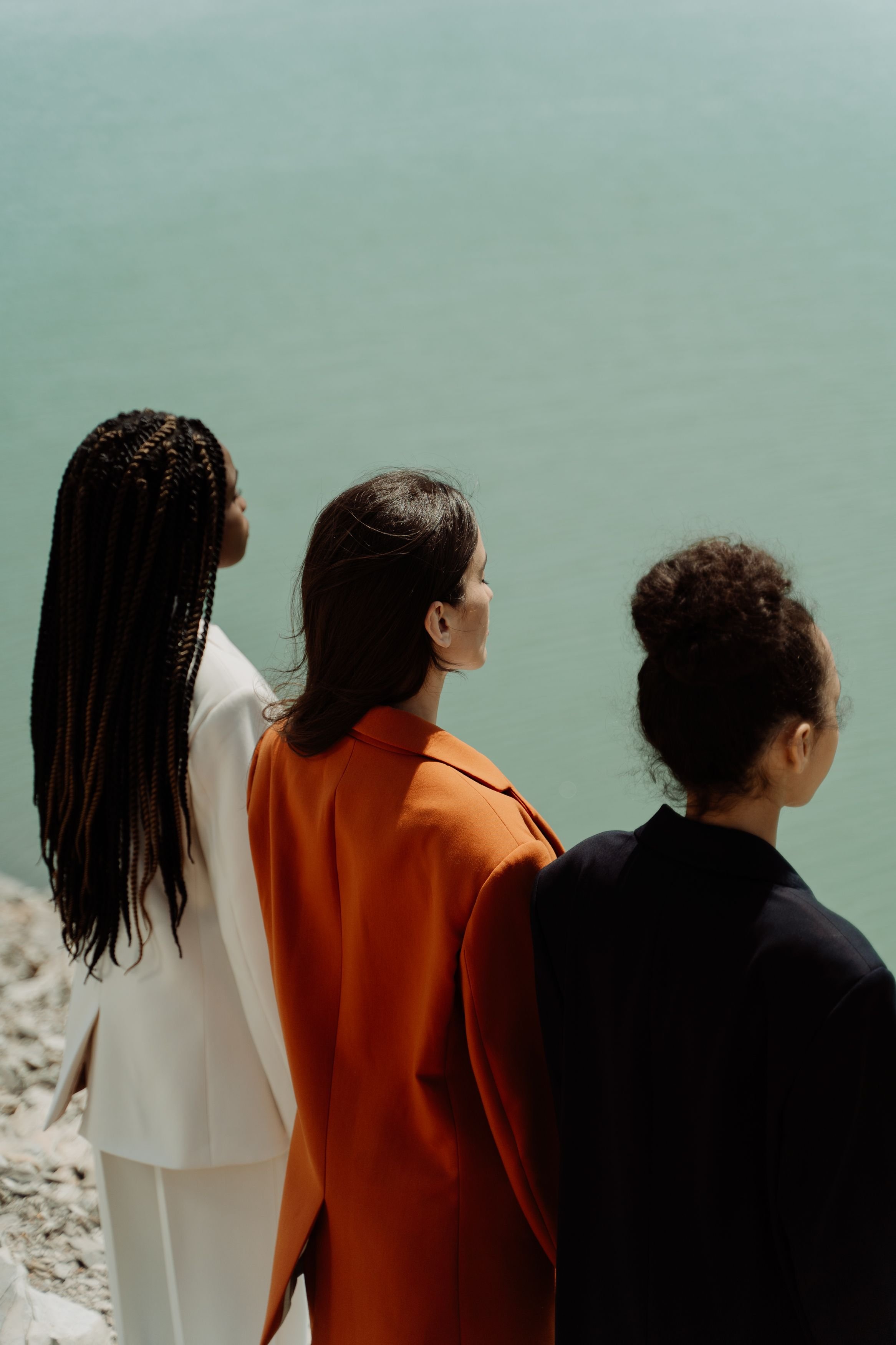 Four women standing on rocky ground near water, facing to the right, with one woman in white on the far left, a woman in orange next, then a woman in black, and a woman in dark brown on the far right.