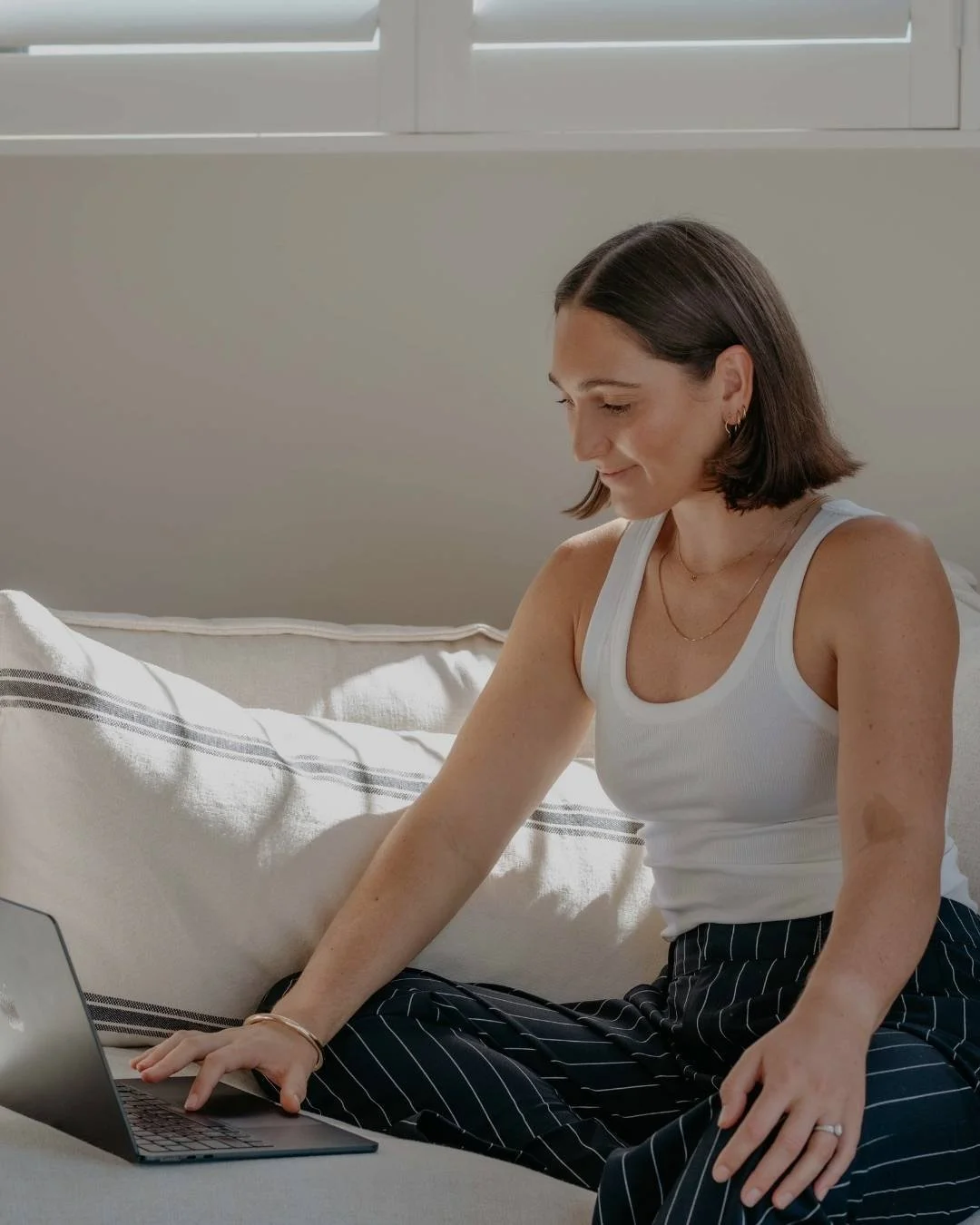 A woman with shoulder-length brown hair, wearing a white tank top and black pinstripe pants, sitting on a beige sofa, using a laptop, and smiling while looking at the screen.