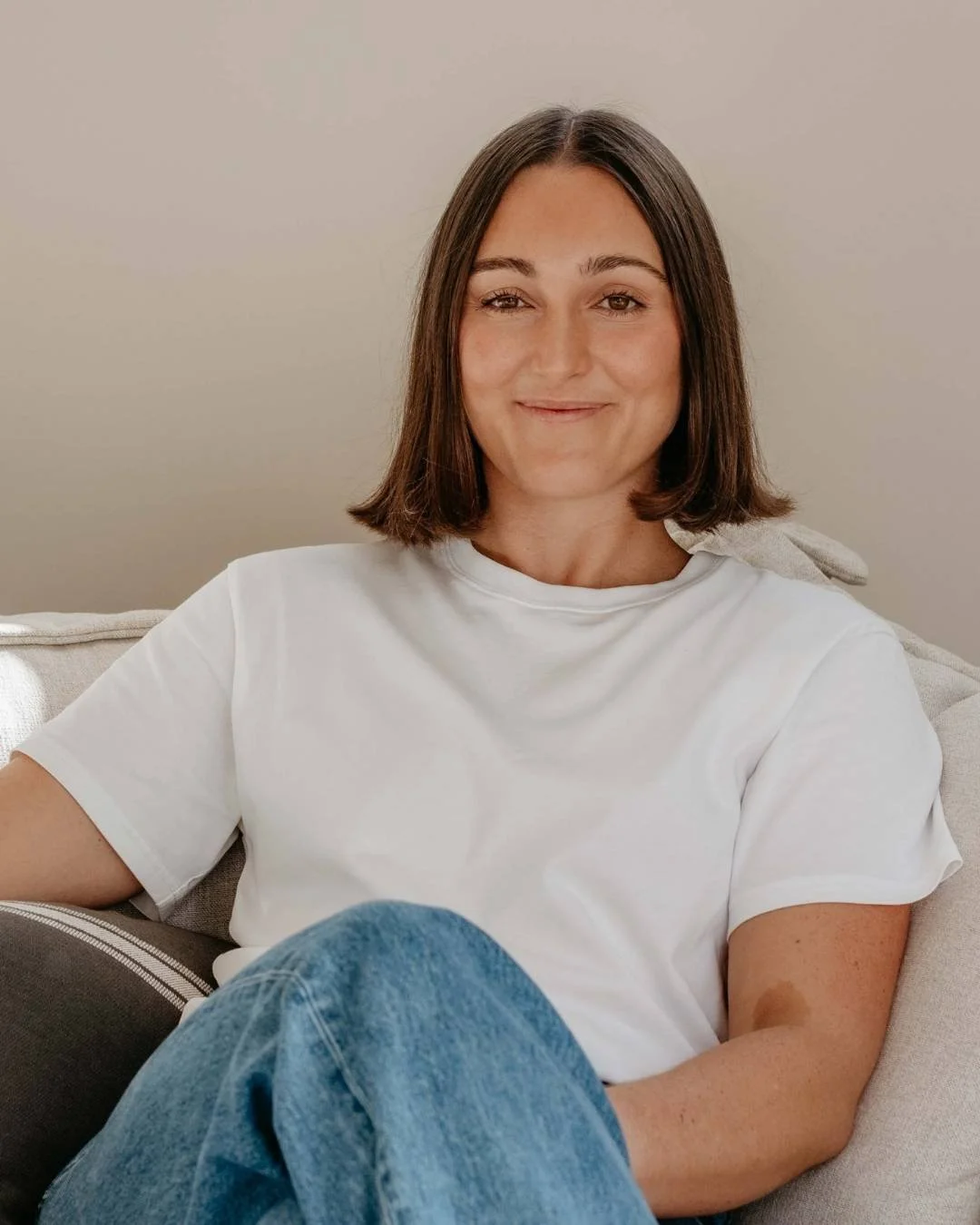 A woman with shoulder-length brown hair, wearing a white t-shirt and blue jeans, sitting on a light-colored couch and smiling at the camera.