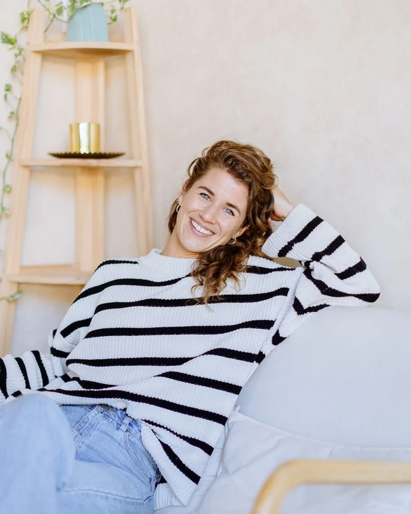A woman with curly red hair smiling and relaxing on a white sofa in a cozy, well-lit room.