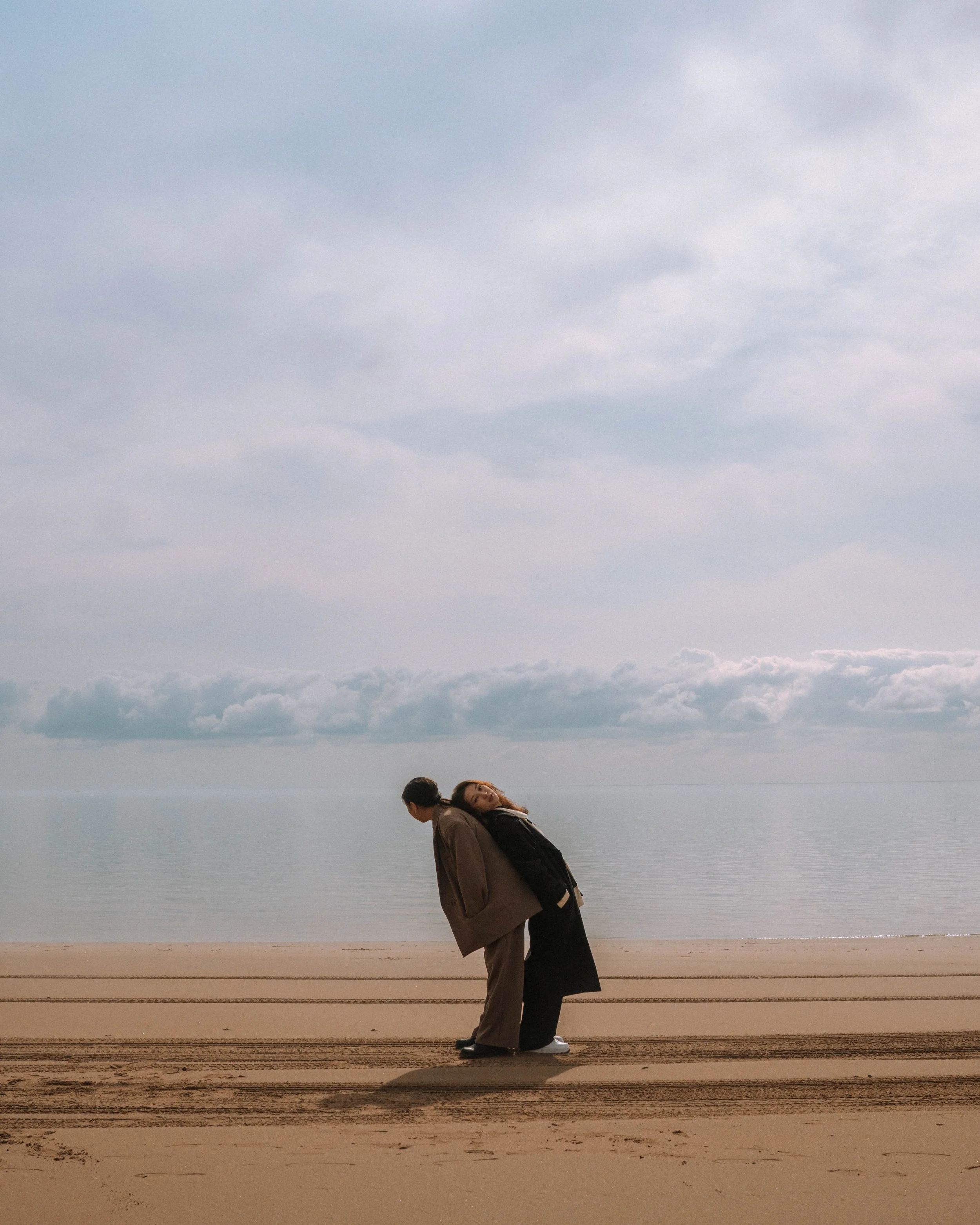 A couple standing close together on a sandy beach with the ocean and cloudy sky in the background. The woman is leaning her head on the man's shoulder, and they appear to be relaxing.
