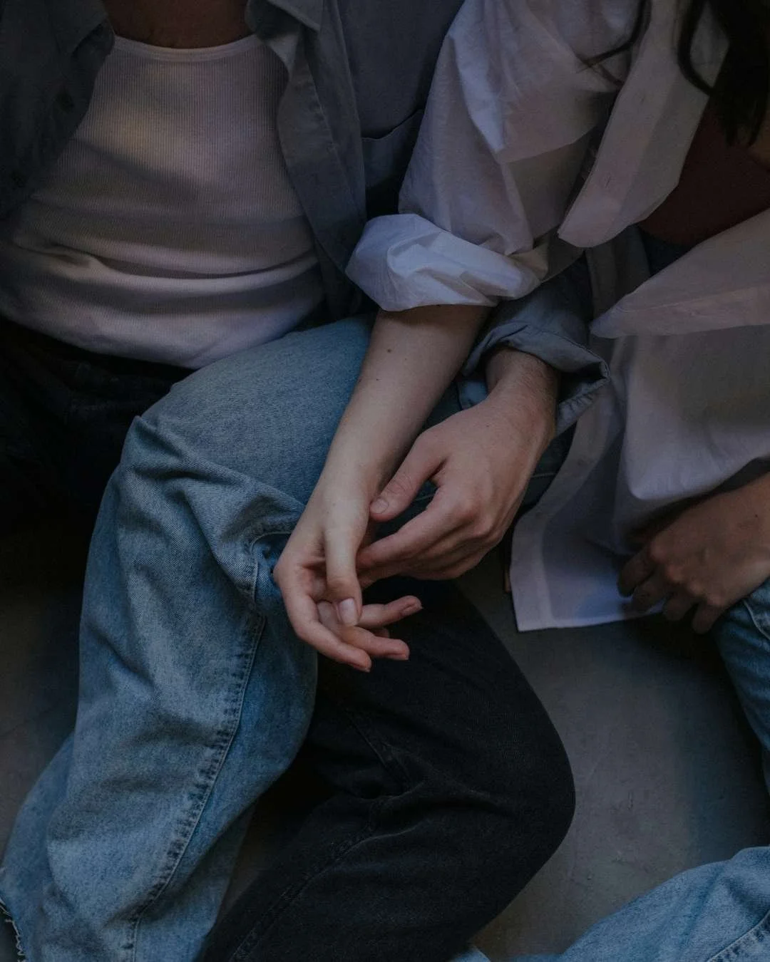 Two people sitting close together, one wearing a white ribbed shirt and jeans, the other in a gray t-shirt and a light-colored jacket, with hands intertwined.