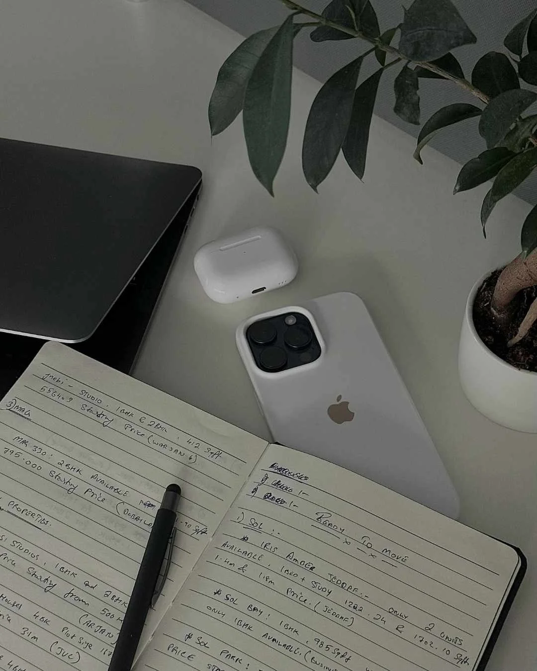Desk with a MacBook, wireless earbuds case, iPhone with a triple-lens camera, open notebook with handwritten notes, pen, potted plant, and part of a laptop.