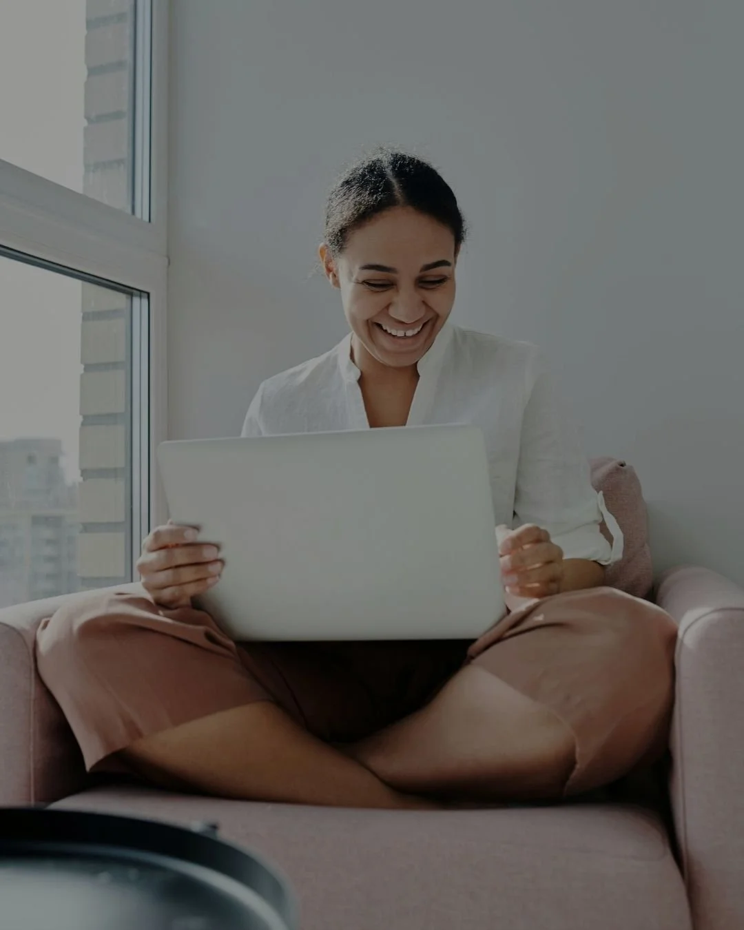 A woman sitting on a pink couch, smiling while looking at her laptop, in a bright room with large window and city buildings outside.