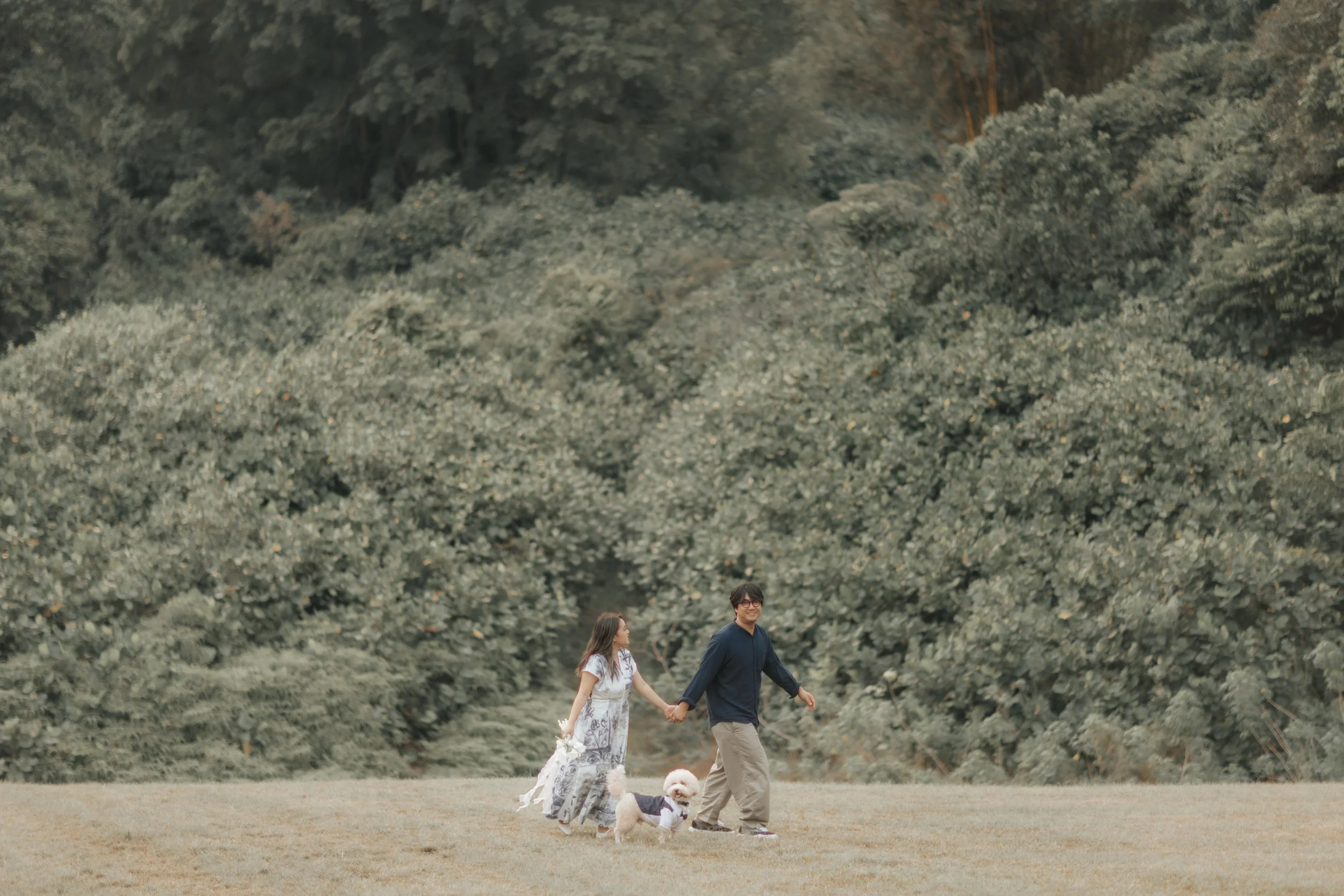 Outdoor pre-wedding photo of a couple walking hand in hand with their dog in a lush green field, captured by Breadandbutterworks in a soft and heartfelt style.