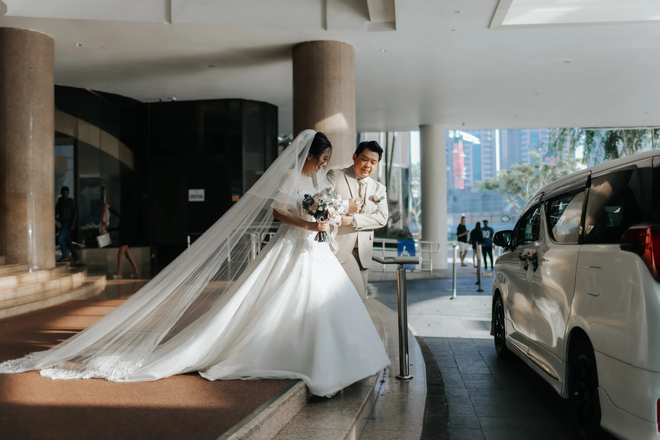 Cinematic actual day wedding photoshoot of a groom ushering his bride-to-be towards their matrimonial car. The shot captures the bride's veil and gown trialing gracefully behind her. Captured in a light and airy style by Arielle for Breadandbutterwor