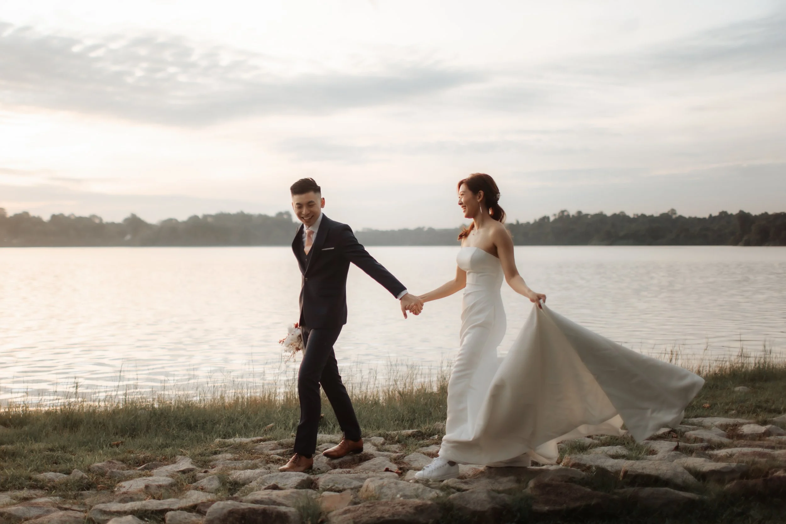 Romantic pre-wedding photoshoot at Upper Seletar Reservoir during golden hour sunset, the couple strolls along the water's edge. Captured in the light and airy style of Breadandbutterworks.