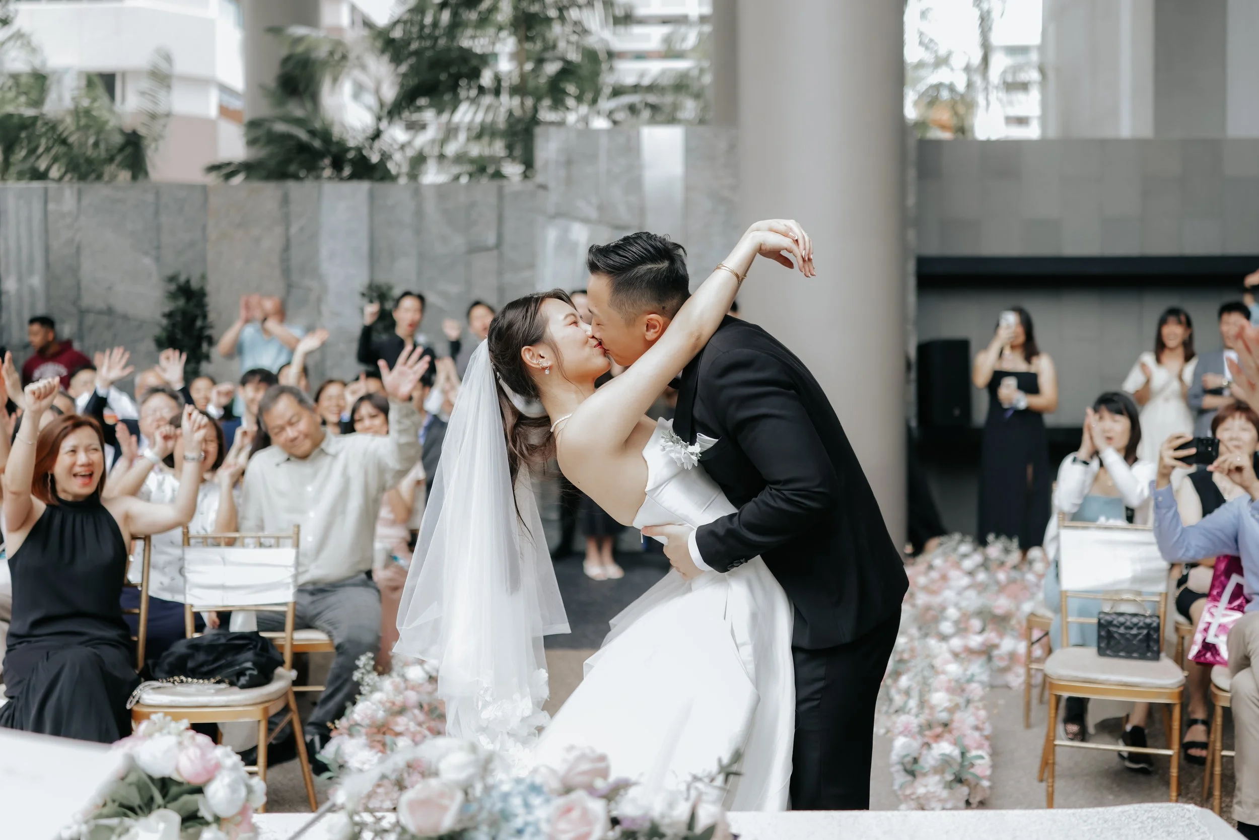 Romantic actual day wedding photography of the couple doing their wedding kiss to the cheers and shouts of their guests, captured by Breadandbutterworks in a soft and heartfelt style.