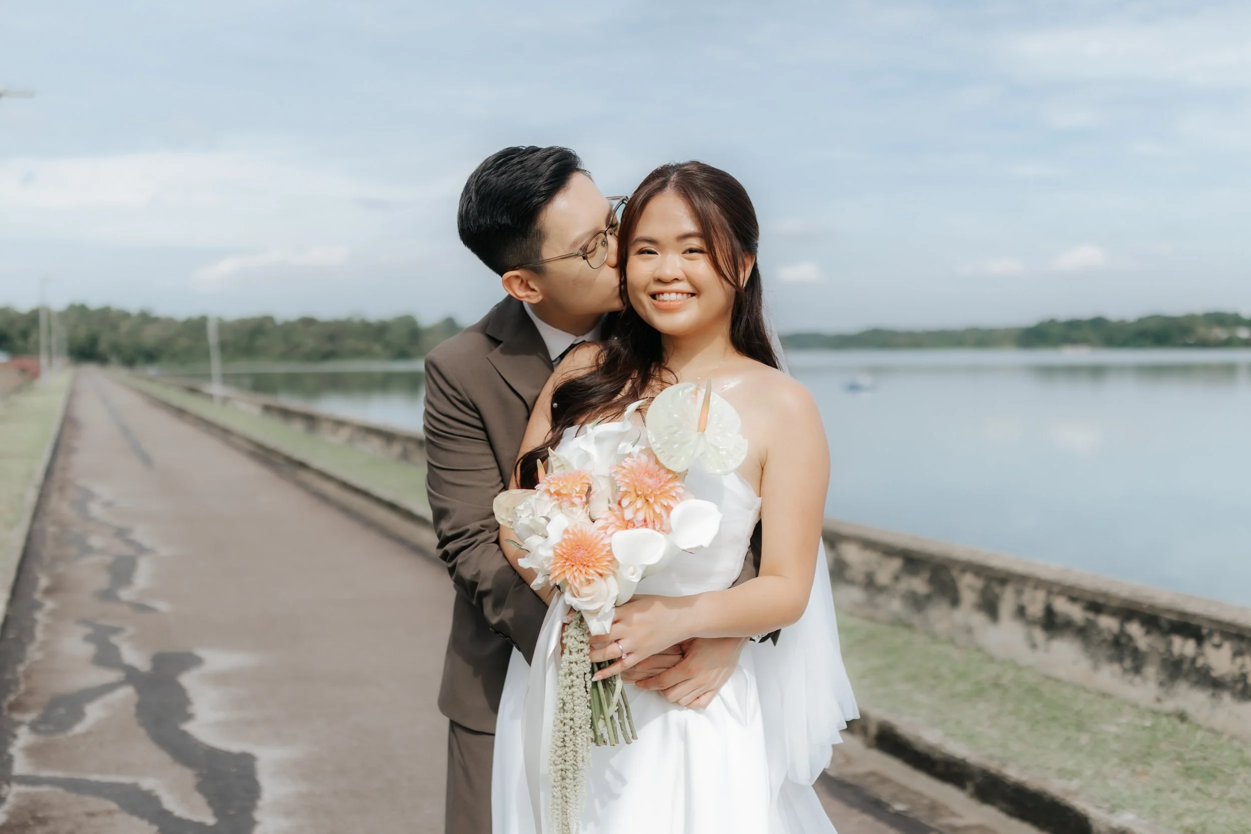 The wedded couple pose for a couple photo with their wedding bouquet beside a body of water. The light and airy vibes was captured by Breadandbutterworks.
