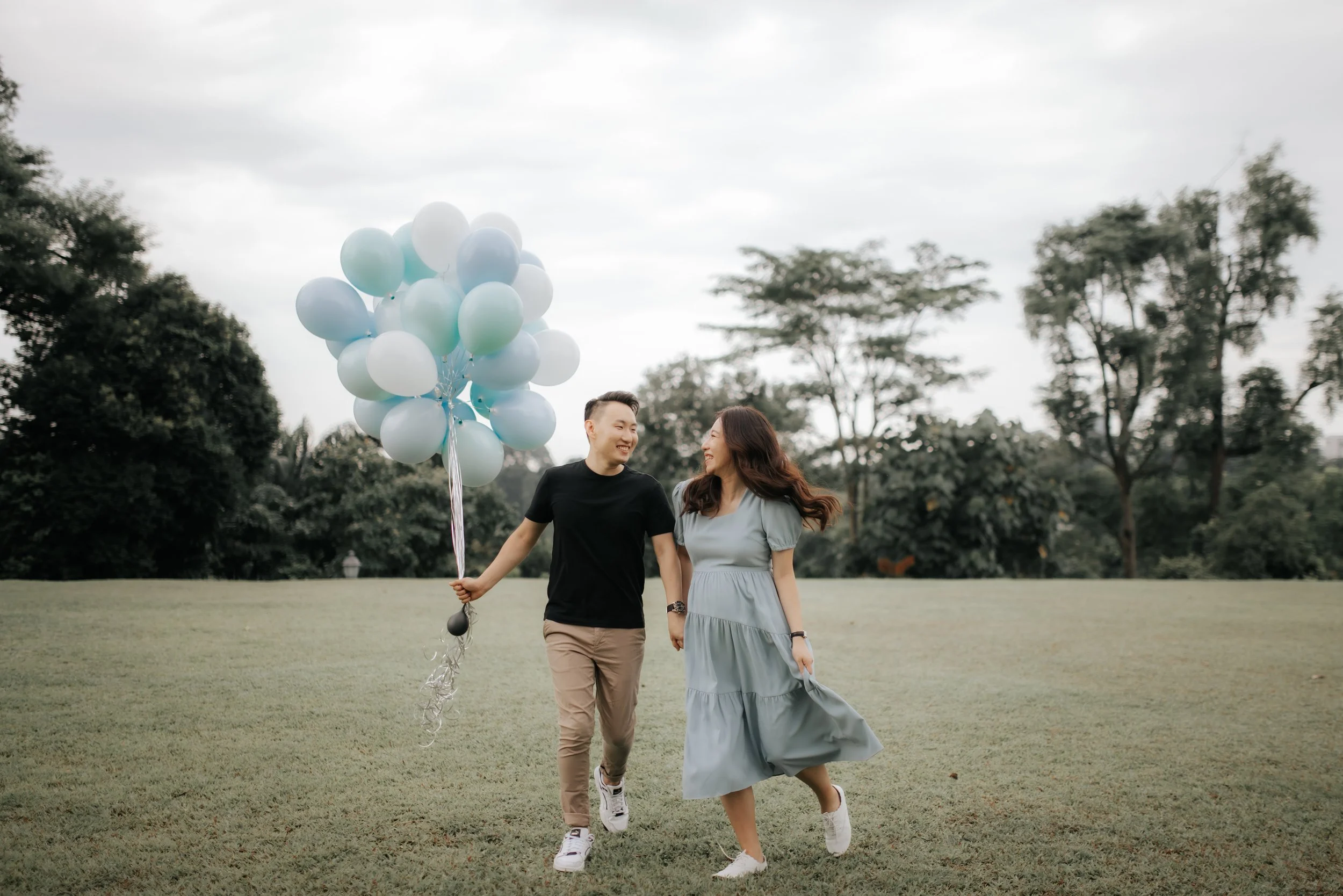 A couple during their maternity shoot taking a stroll at Gallop Extension in Singapore while holding on to a bunch of balloons, captured by Breadandbutterworks in a soft and heartfelt style.
