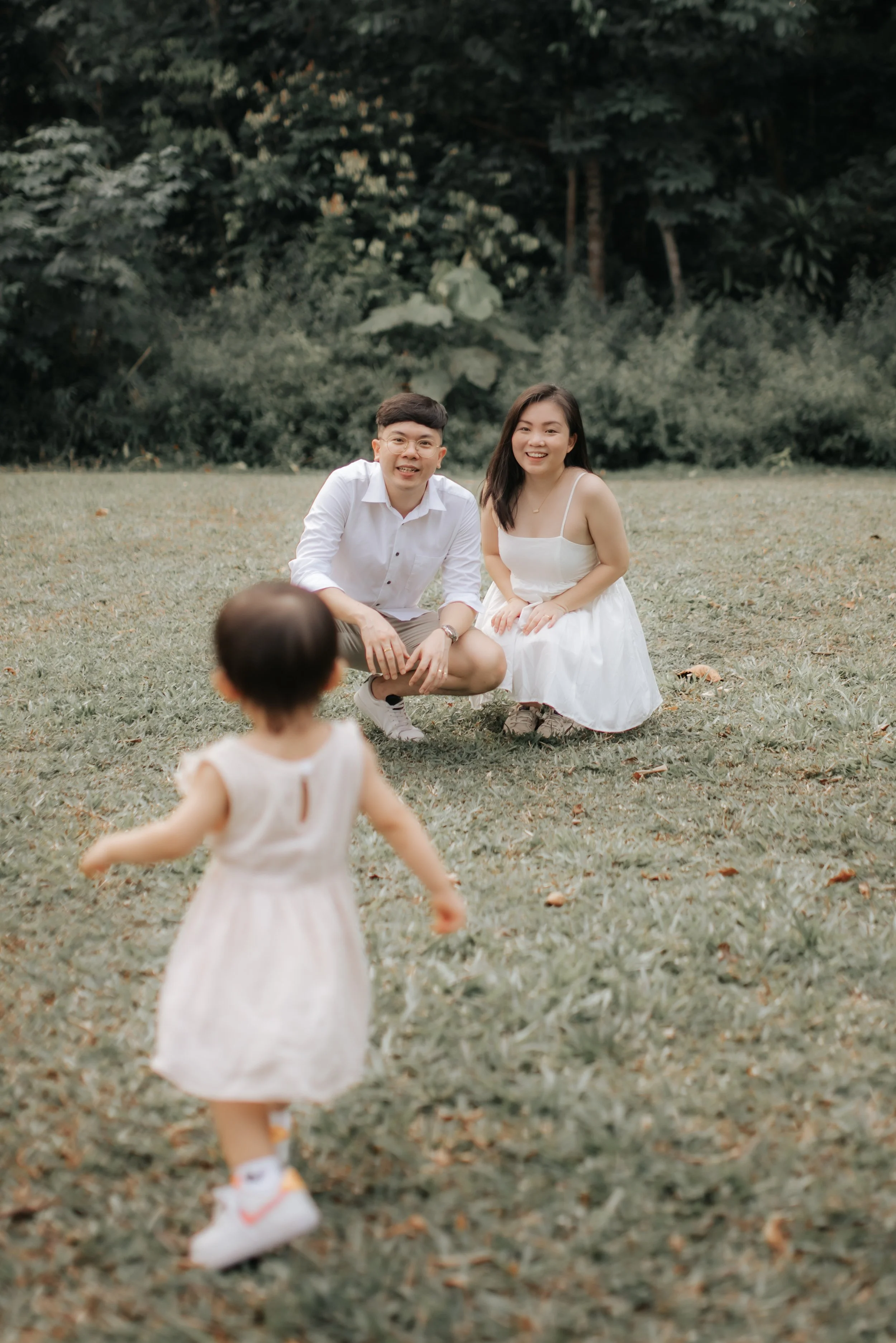 Light and airy family photo of a young family encouraging their child as she runs towards them, captured in a soft outdoor setting in Singapore.