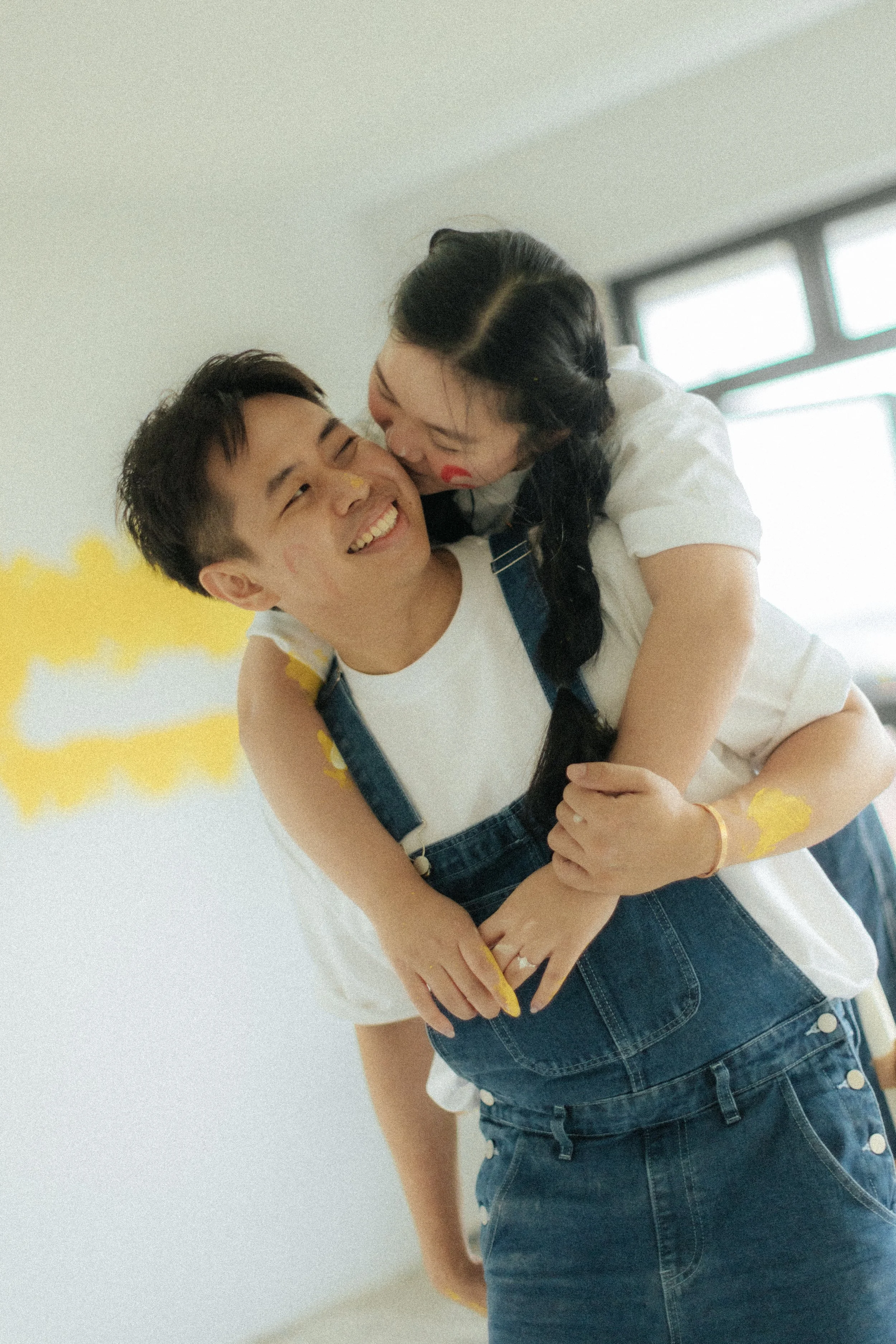Playful home pre-wedding photo of a couple painting their marital home together, they share a moment with a back hug. Captured by Breadandbutterworks in a light, airy and heartfelt style.