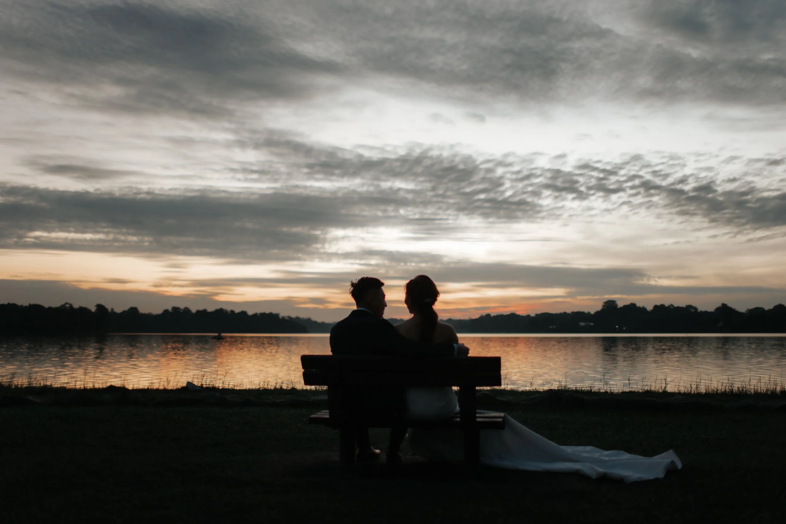 Creative romantic pre-wedding photoshoot at Upper Seletar Reservoir during golden hour sunset, the couple forms a darling silhouette while sitting on a bench watching the sunset. Captured in the light and airy style of Breadandbutterworks.