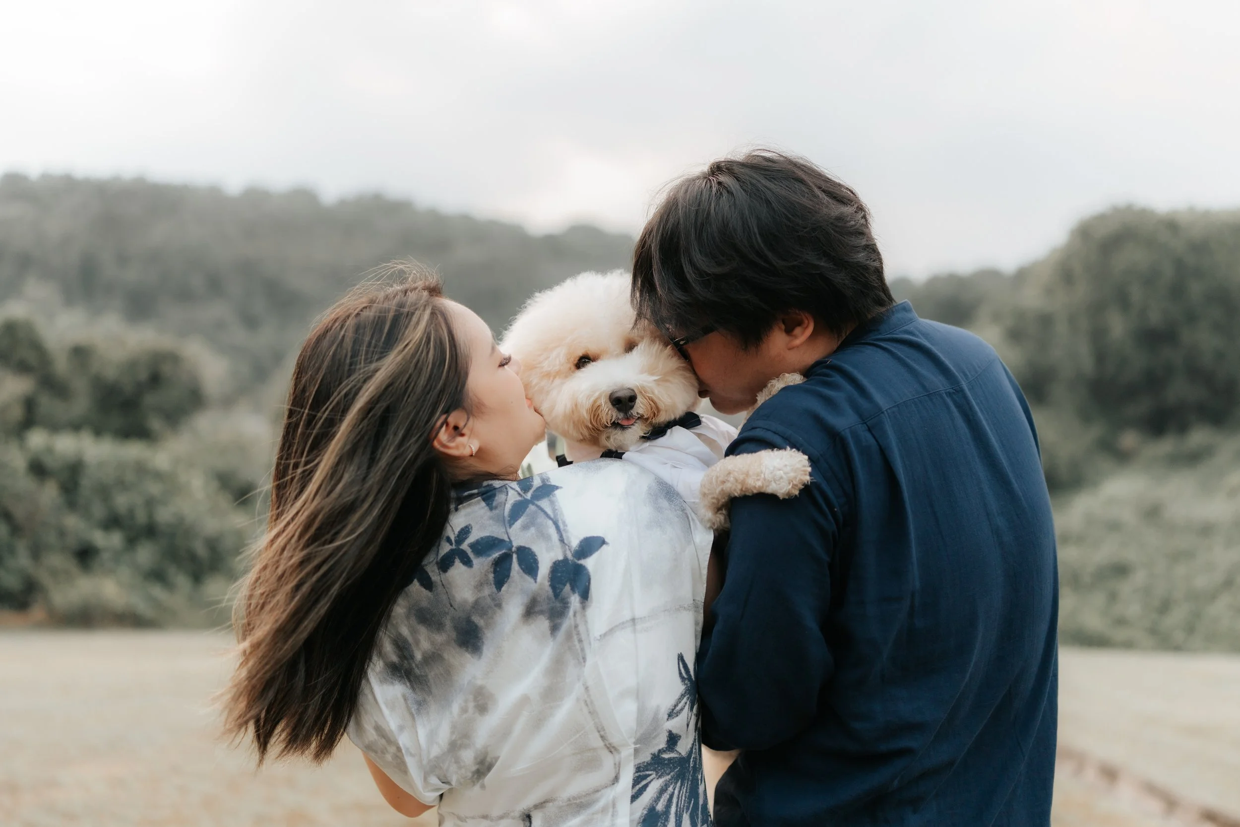 Outdoor pre-wedding photo of a couple cuddling with their dog in a lush green field in Canterbury, captured by Breadandbutterworks in a soft and heartfelt style.