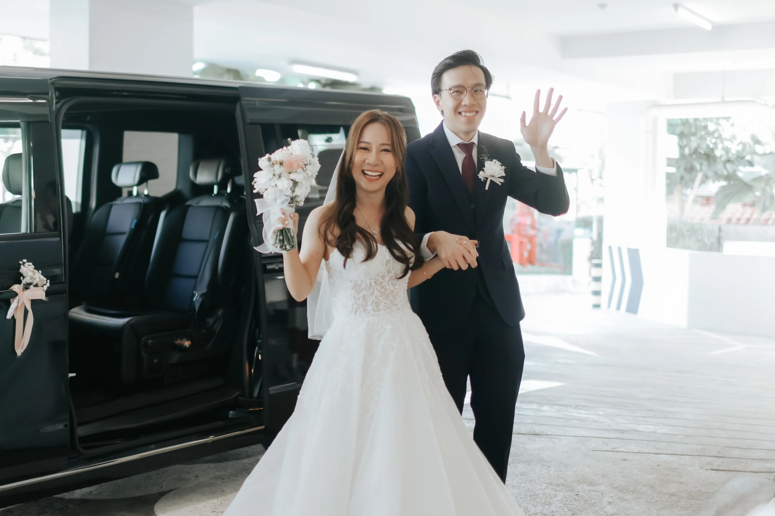Light and airy actual wedding day photoshoot of the bride and groom emerging from their matrimonial car as they wave towards the camera with happy smiles. Captured by Arielle for Breadandbutterworks.