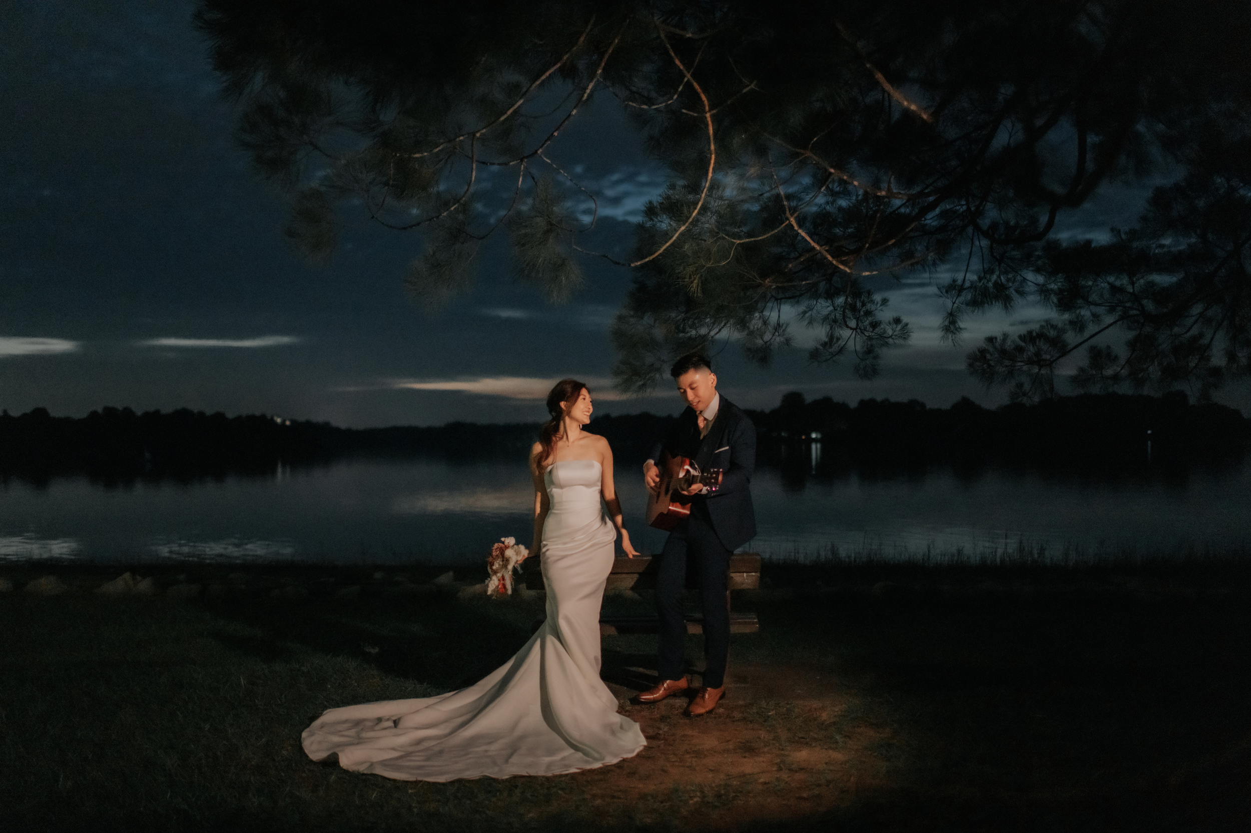 Romantic pre-wedding night photoshoot at Upper Seletar Reservoir, the groom serenades the bride with his guitar and song. There is a cinematic lighting effect in this photo achieved through the use of a spotlight. Captured in the light and airy style