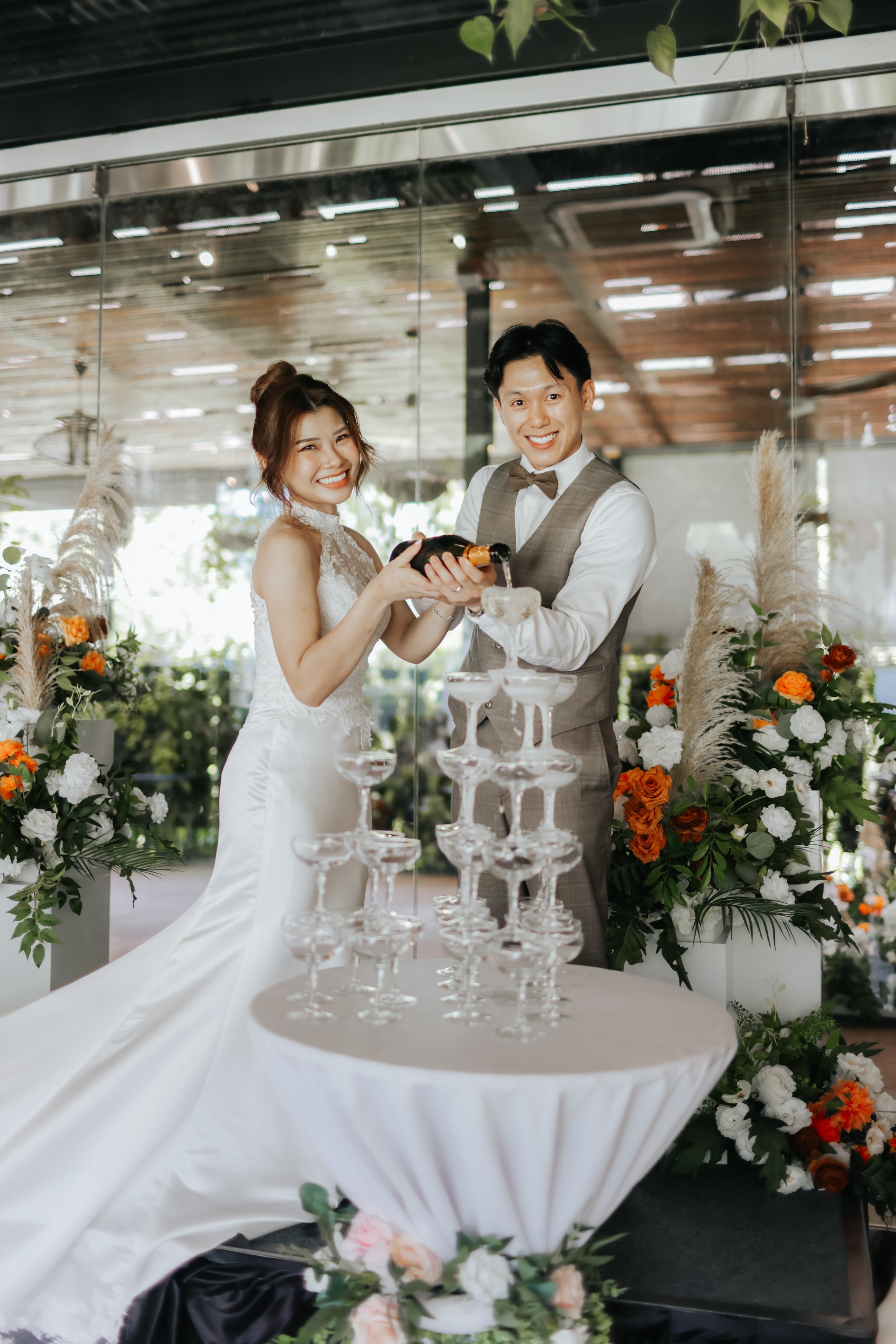 Light and airy actual day wedding photoshoot at Canopy HortPark, the couple pours champagne over the tower of glasses. Captured by Arielle from Breadandbutterworks.