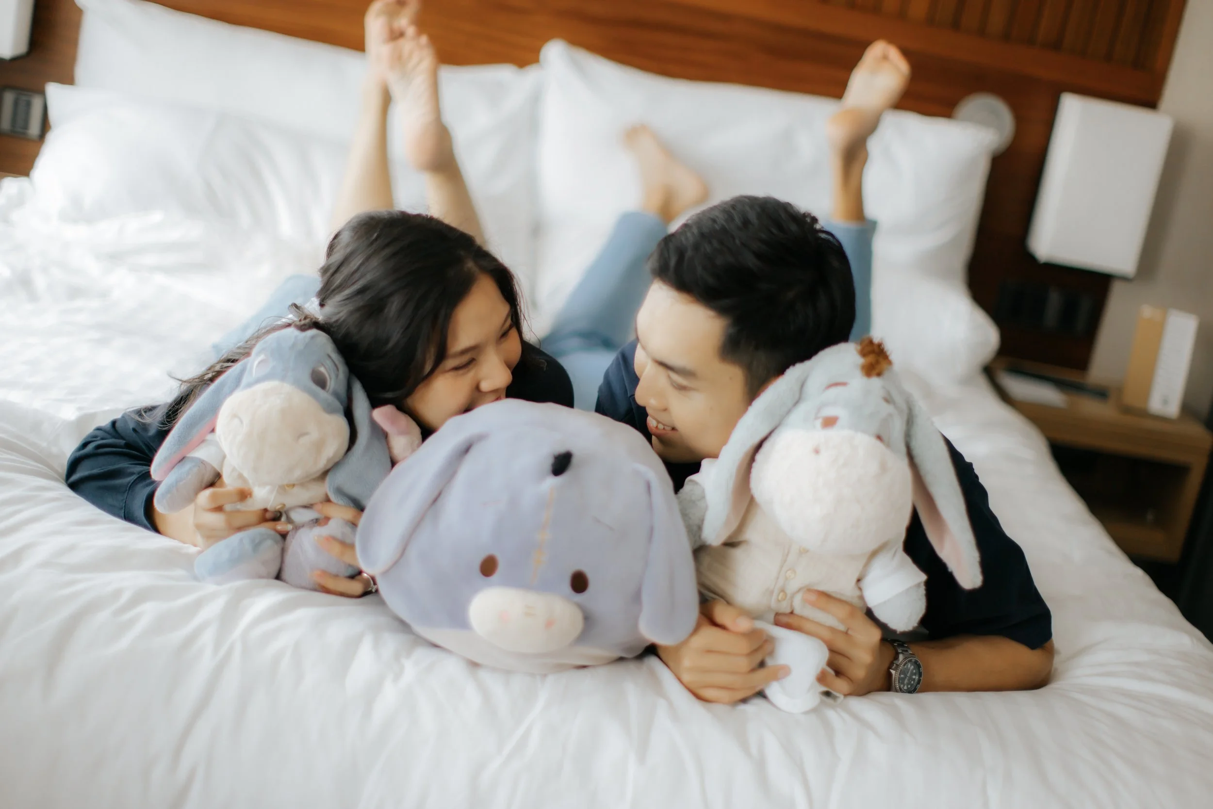 Cosy home pre-wedding photo of a couple lying on a bed with plush toys, captured by Ash for Breadandbutterworks in a light, airy and heartfelt style.