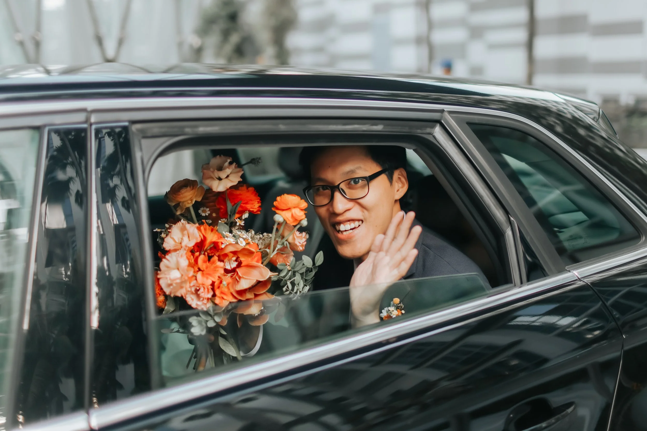 Actual day wedding photoshoot of a groom arriving in the matrimonial car with a autumn coloured bouquet in hand, he waves cheerfully at the camera. Shot in a light and airy style by Arielle for Breadandbutterworks.