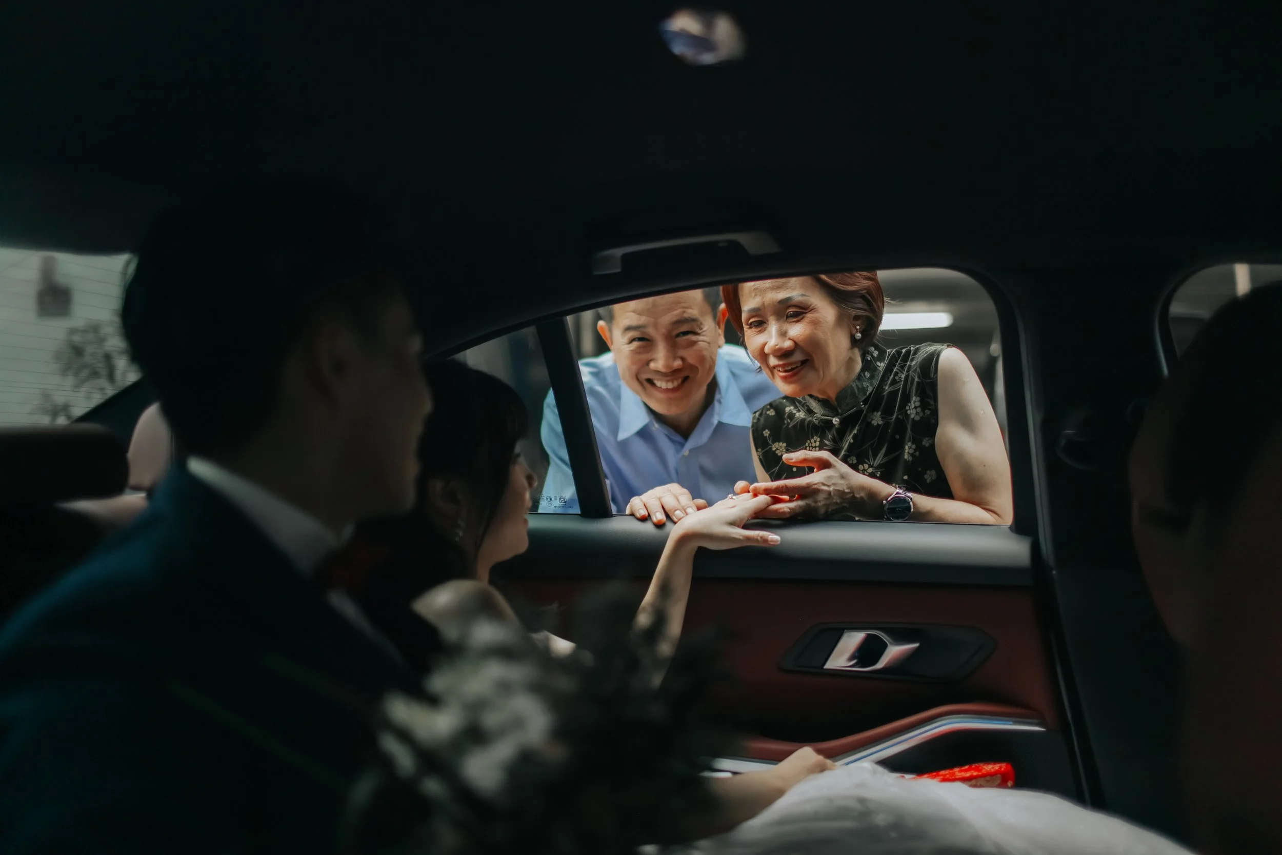 A bride and groom on their actual wedding day sitting in their matrimonial car, with the bride's parents cinematically captured through the car window. Captured by Arielle for Breadandbutterworks.