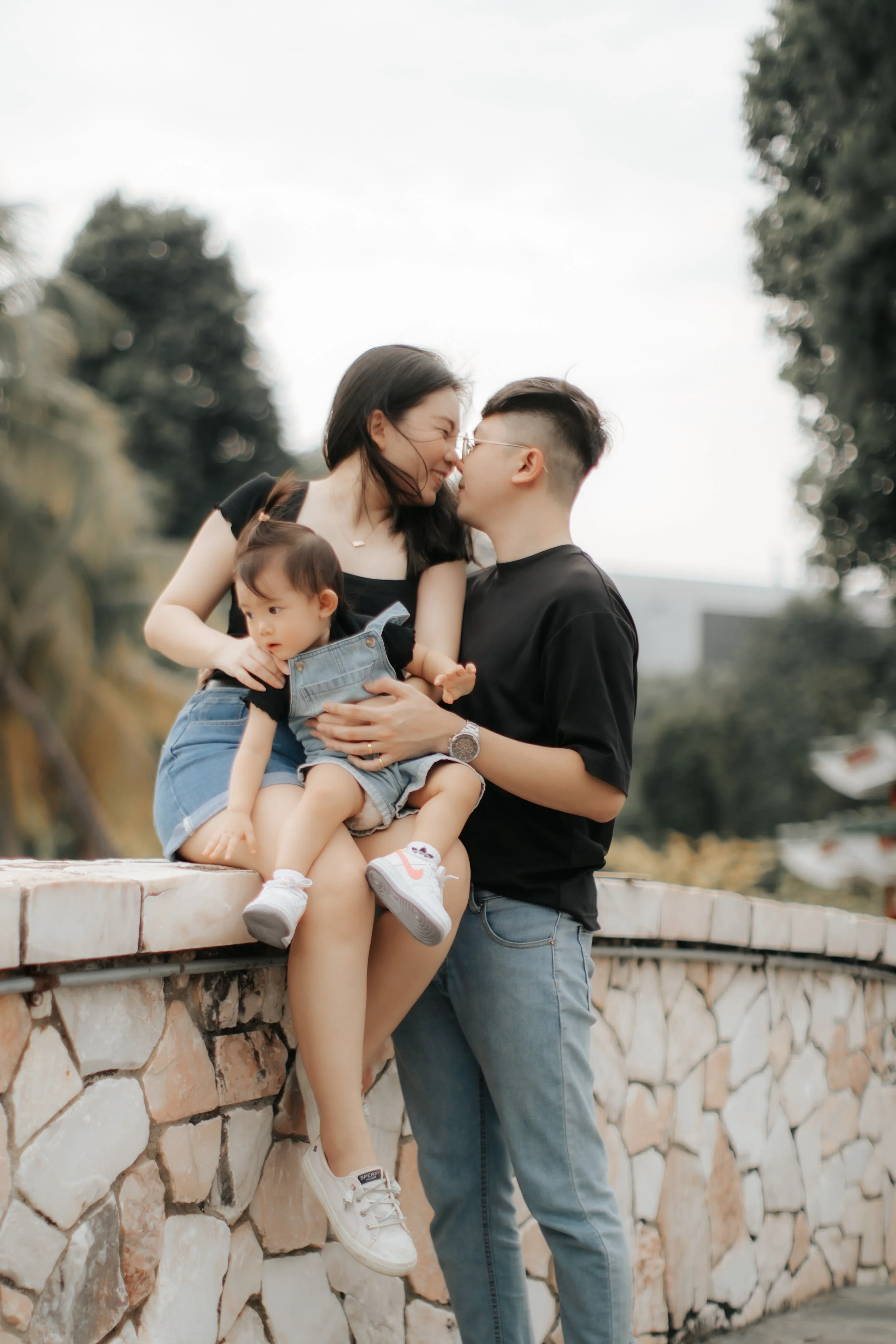 Light and airy family photo of a young family sharing a sweet moment on a bridge, captured in a soft outdoor setting in Singapore.
