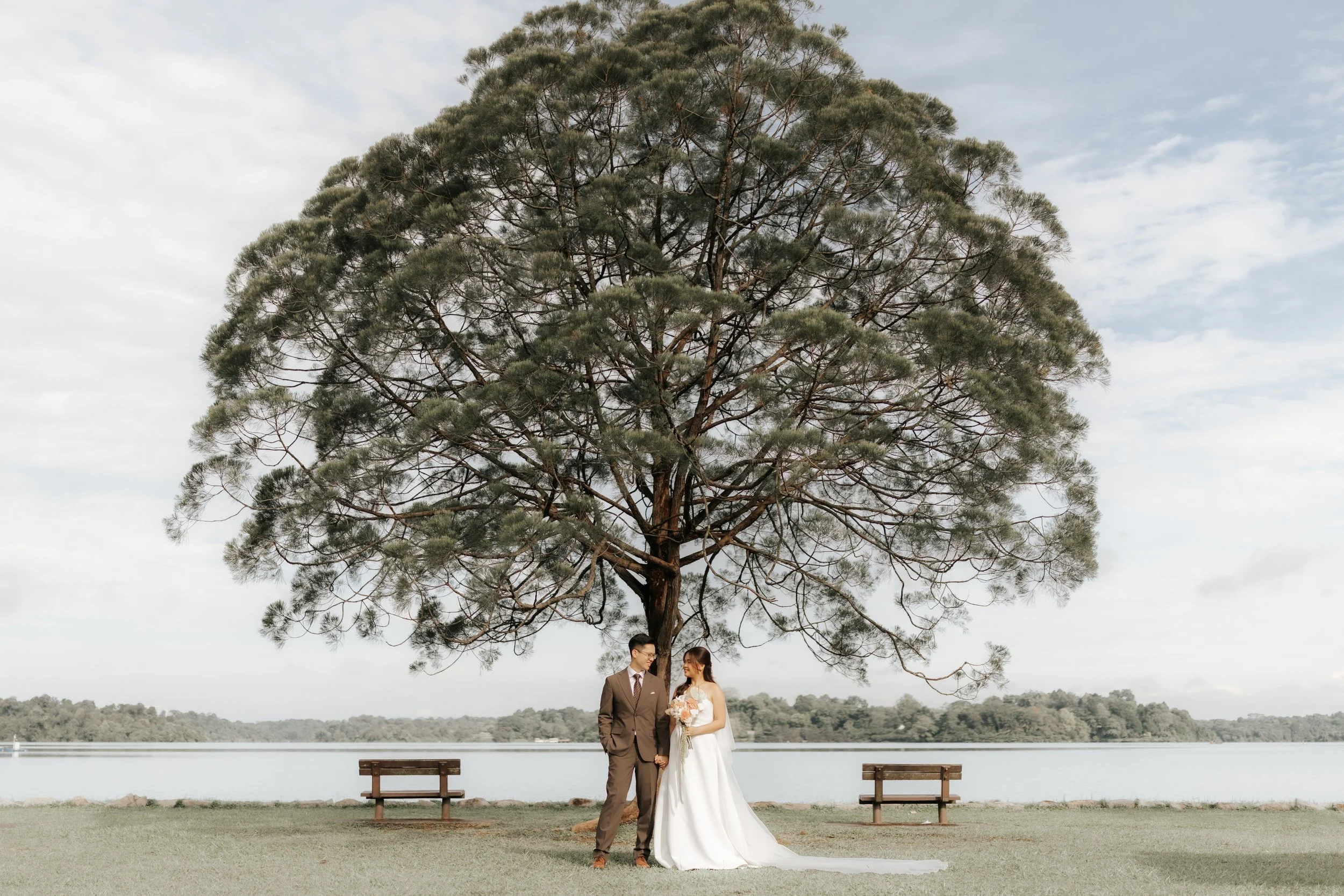 The wedded couple pose for a cinematic photo with a majestic tree and waterfront as their backdrop. The photo was taken by Breadandbutterworks.
