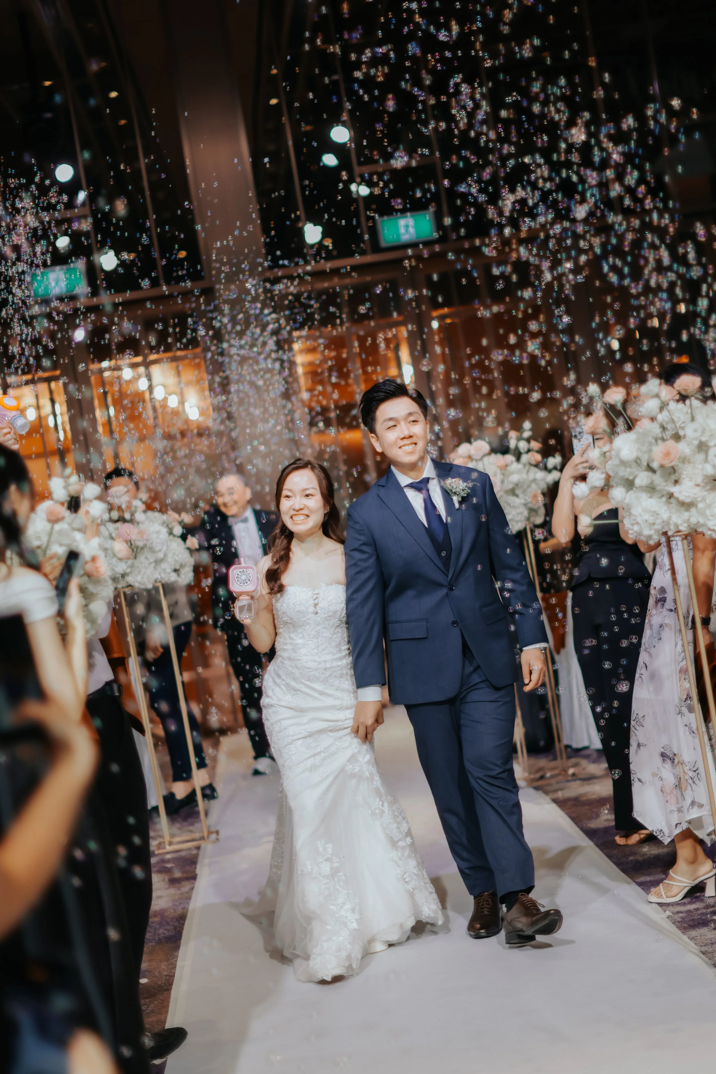Joyful actual day wedding photoshoot of a couple doing their first march in while their friends and loved ones shower them with bubbles, captured by Breadandbutterworks in a soft and heartfelt style.