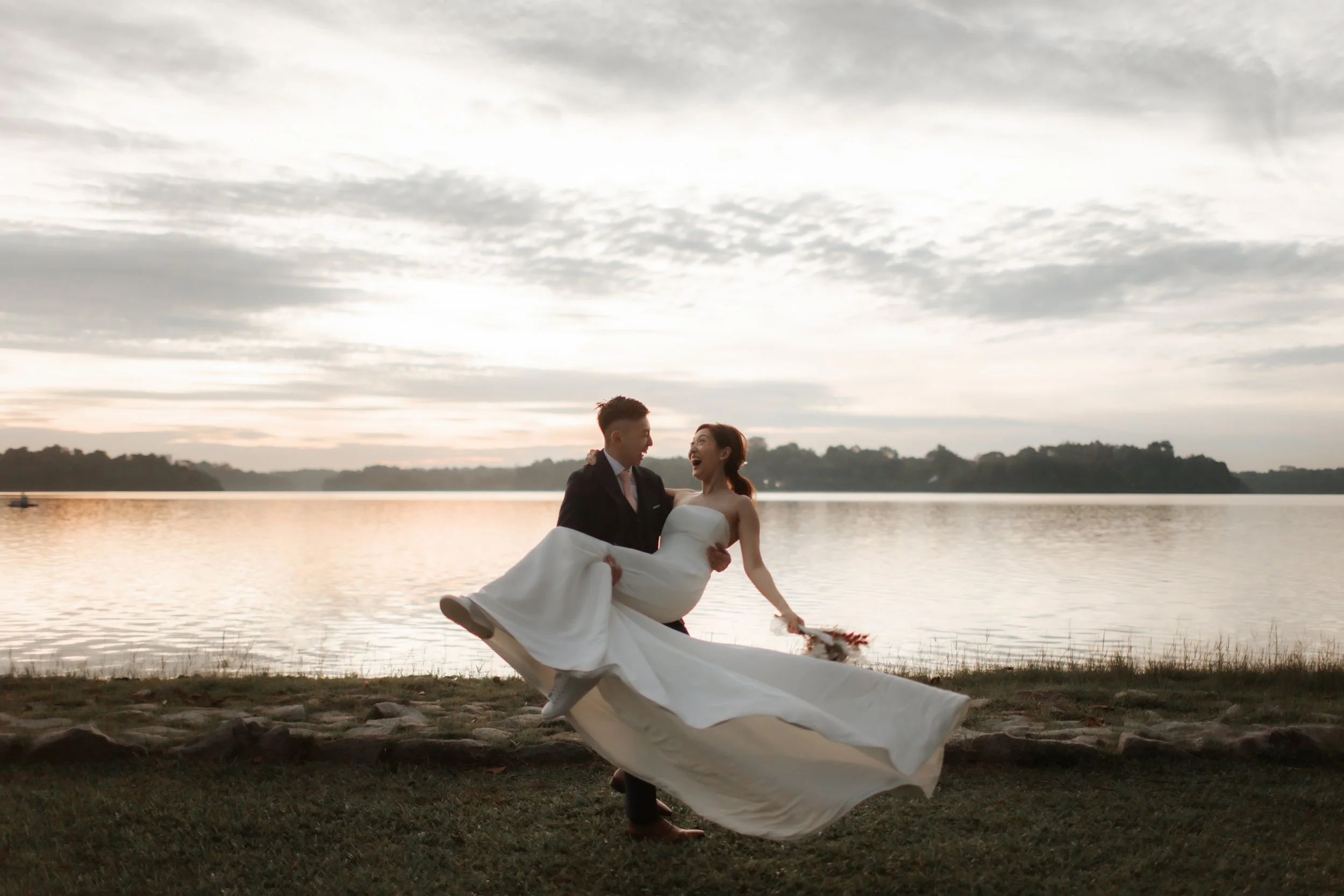 Romantic pre-wedding photoshoot at Upper Seletar Reservoir during golden hour sunset, the groom carries the bride and twirls her around as her gown flows gracefully. Captured in the light and airy style of Breadandbutterworks.