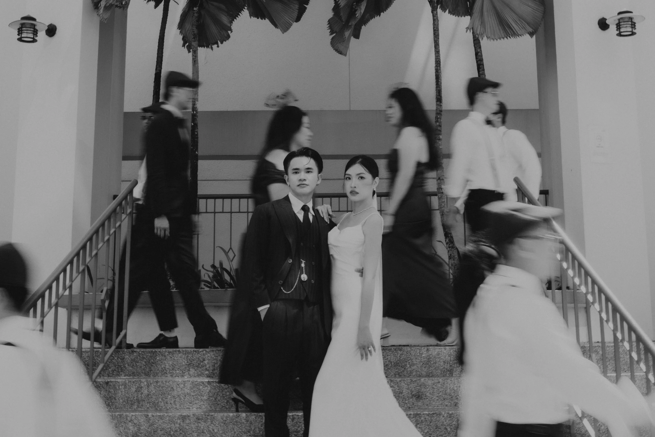 Dynamic black and white actual day wedding photoshoot of the couple and the bridal party. The couple looks on as their friends walk across the frame, forming a juxtaposition of movement and stillness. Captured by Arielle from Breadandbutterworks.