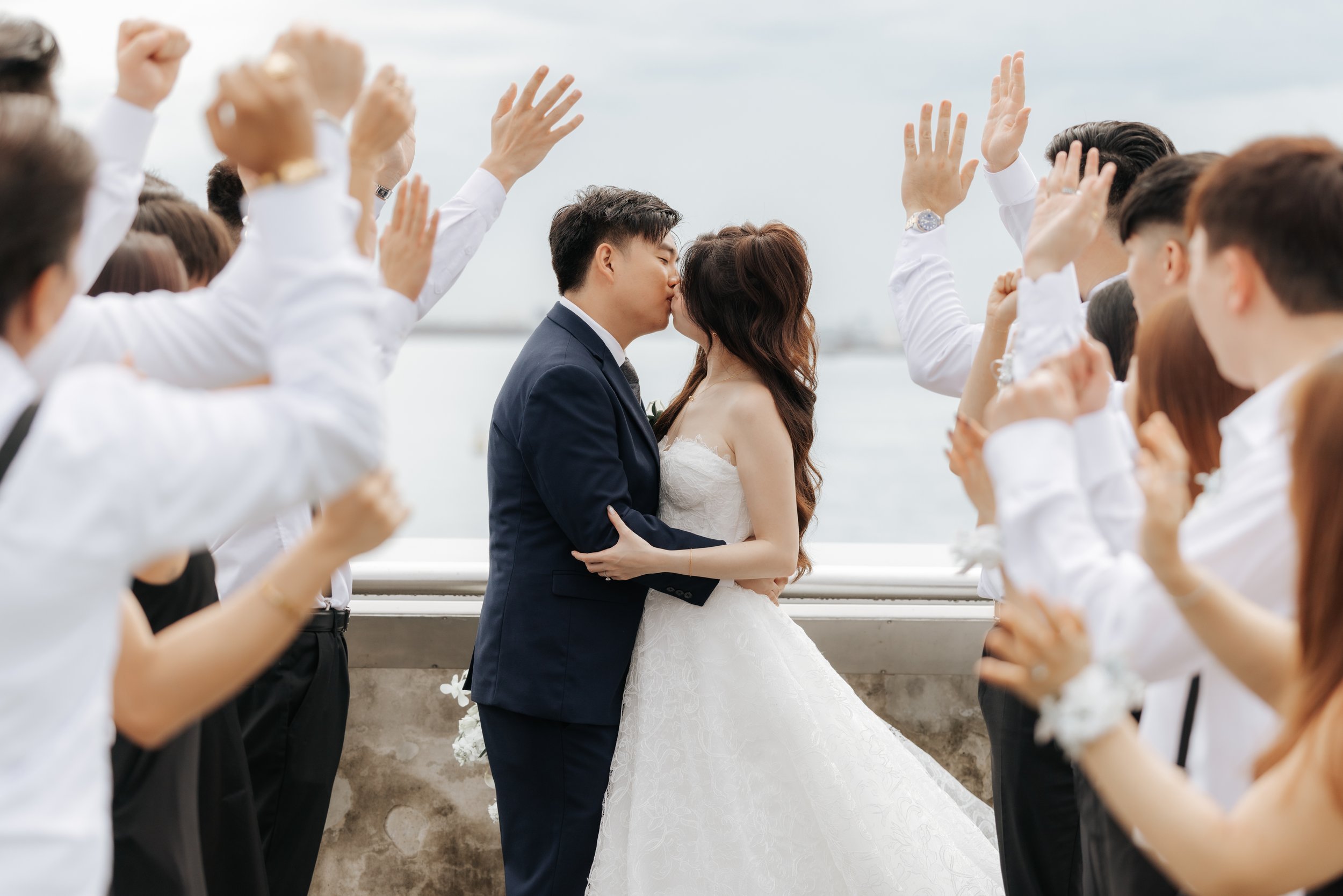 Romantic actual day wedding photo of a couple sharing a kiss while their loved ones cheer them on, this was captured along Marina Barrage by Breadandbutterworks in a soft and heartfelt style.