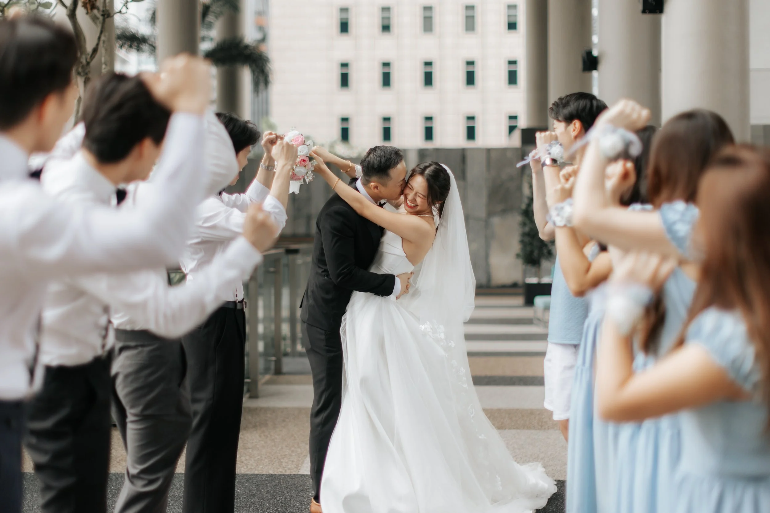 Cheerful and romantic actual day wedding photography of the couple embracing while their bridal party cheers them on, captured by Breadandbutterworks in a soft and heartfelt style.