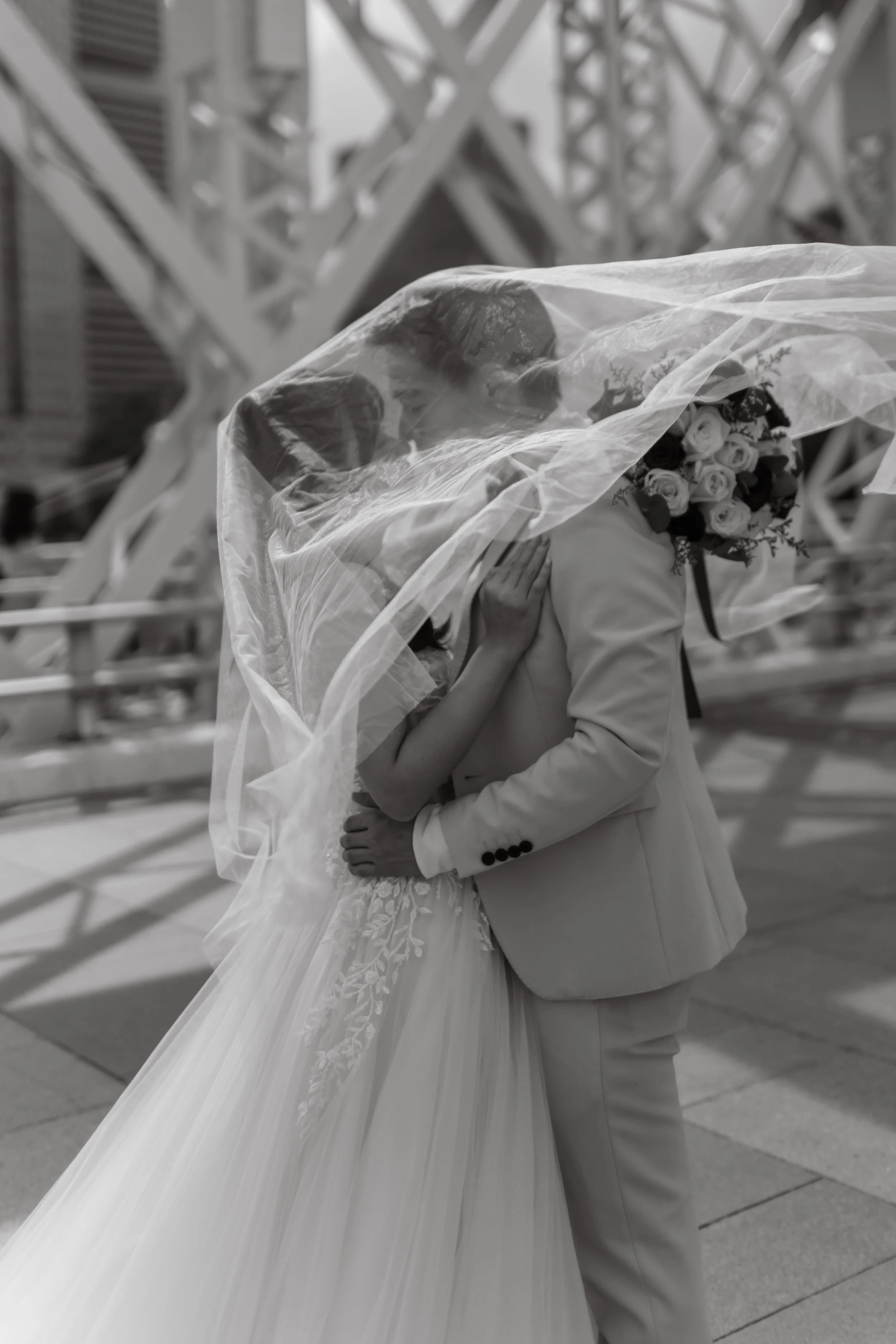 Cinematic and editorial black and white actual day wedding photography of the couple kissing beneath the wedding veil, the veil is being blown gracefully by the wind. Captured by Arielle for Breadandbutterworks.