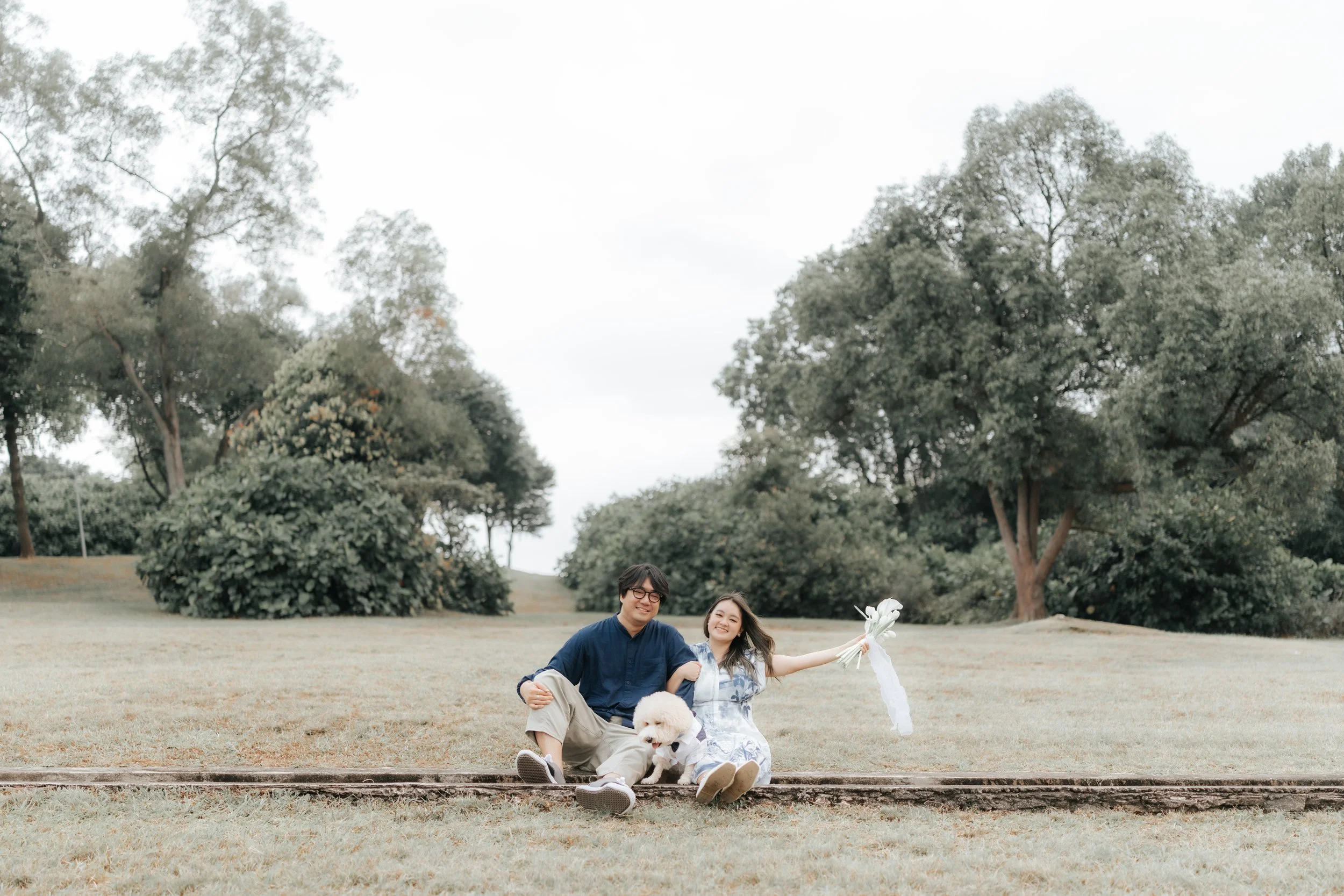 Outdoor pre-wedding photo of a couple hanging out with their dog in a lush green field in Canterbury, captured by Breadandbutterworks in a soft and heartfelt style.
