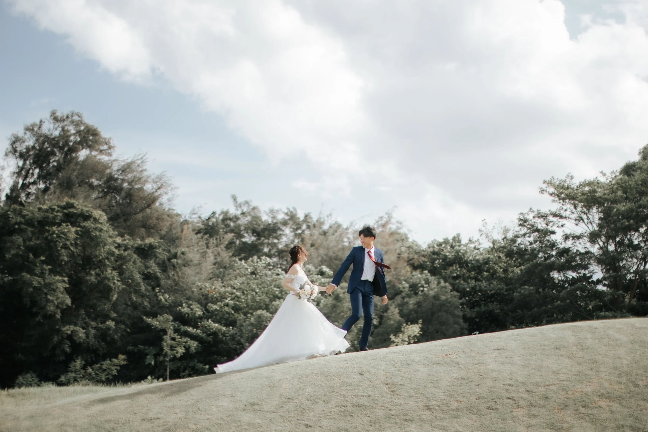 Light and airy pre-wedding photo of a bride and groom walking hand in hand across a grassy hill in Jurong Lake Gardens in Singapore.