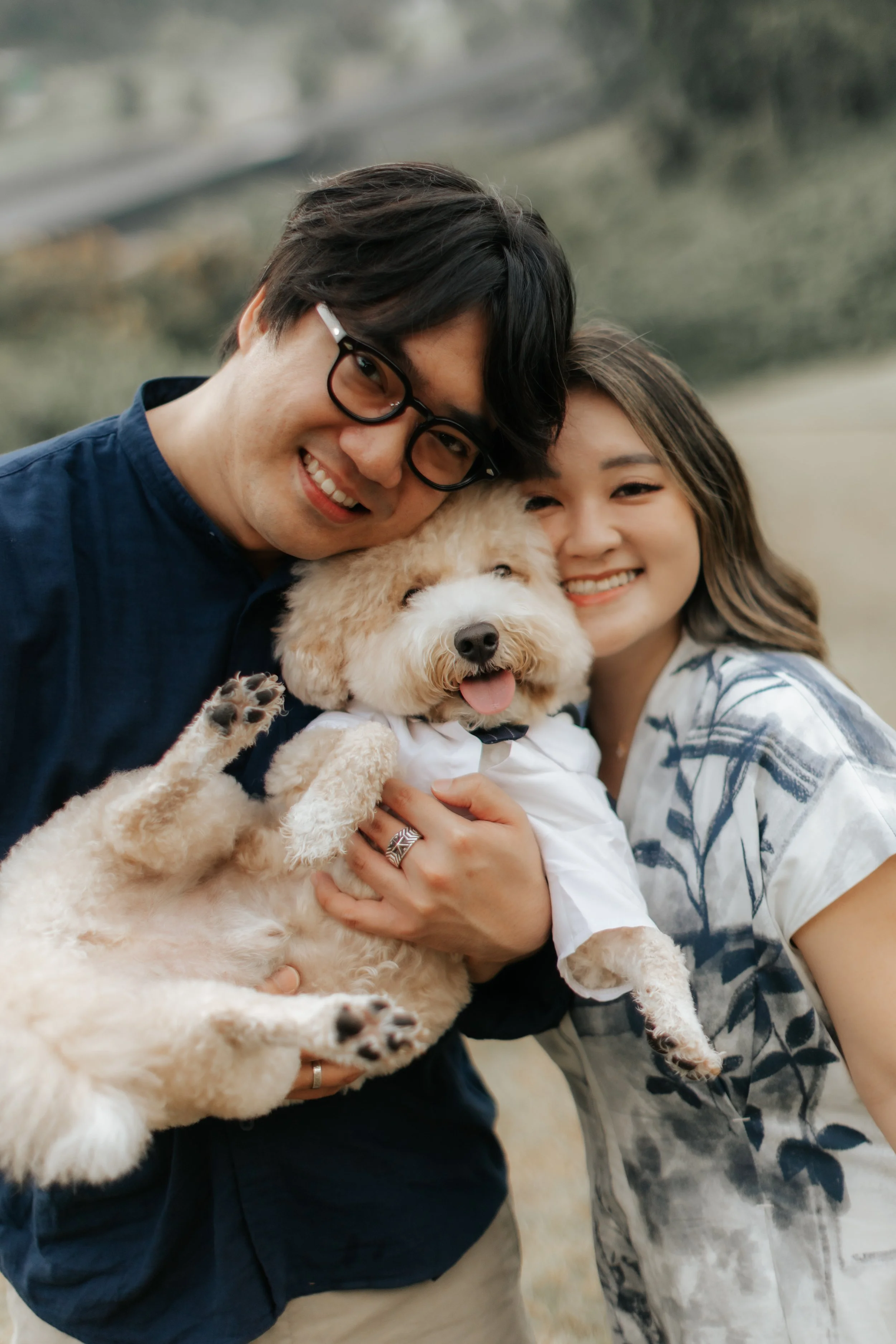 Outdoor pre-wedding photo of a couple posing with their dog in a lush green field, captured by Breadandbutterworks in a soft and heartfelt style.