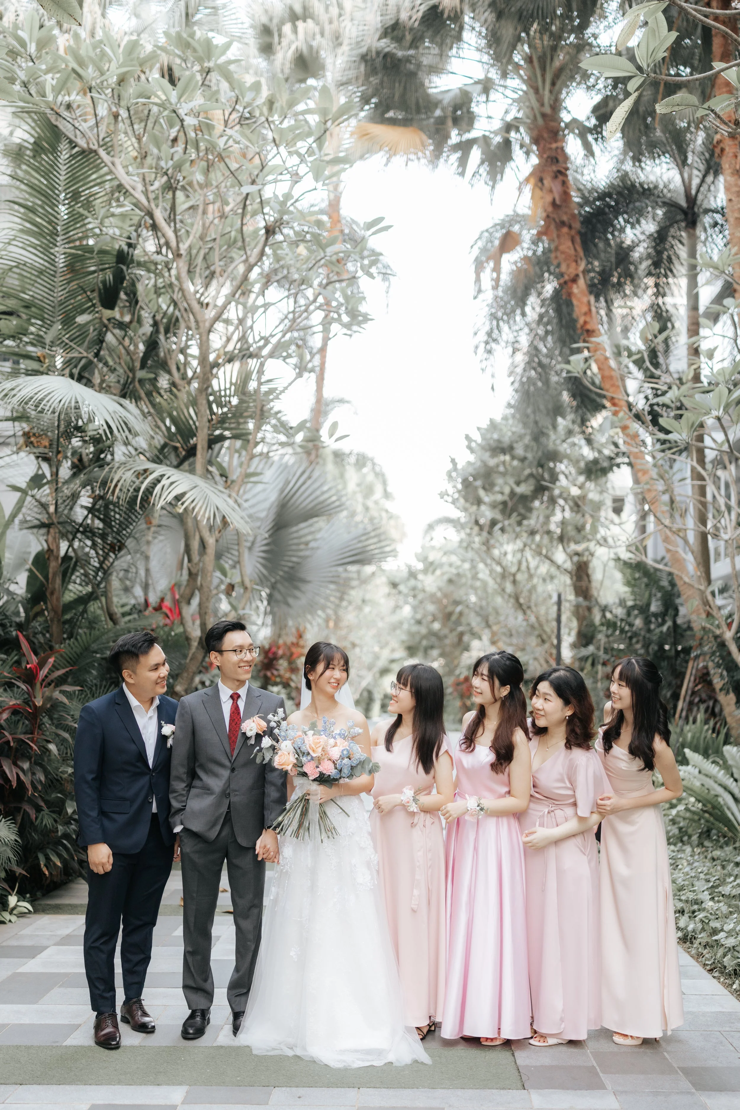 The couple poses with their bridal party along a garden-flanked path, featuring the joyful groomsman and bridesmaids. Photographed by Breadandbutterworks.